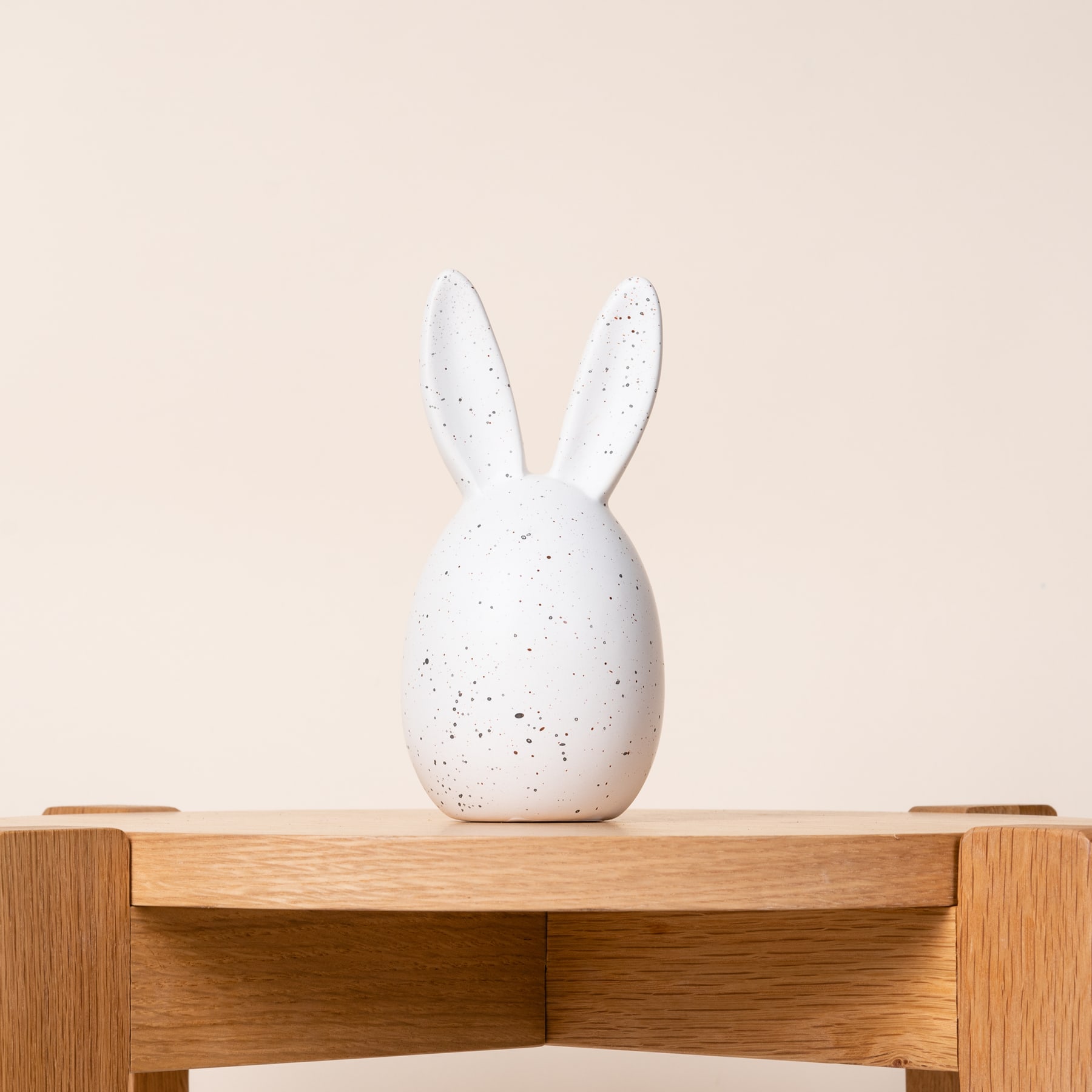 Eine weiße Kaninchenfigur aus Keramik steht auf einem Holzregal vor einem beigen Hintergrund.