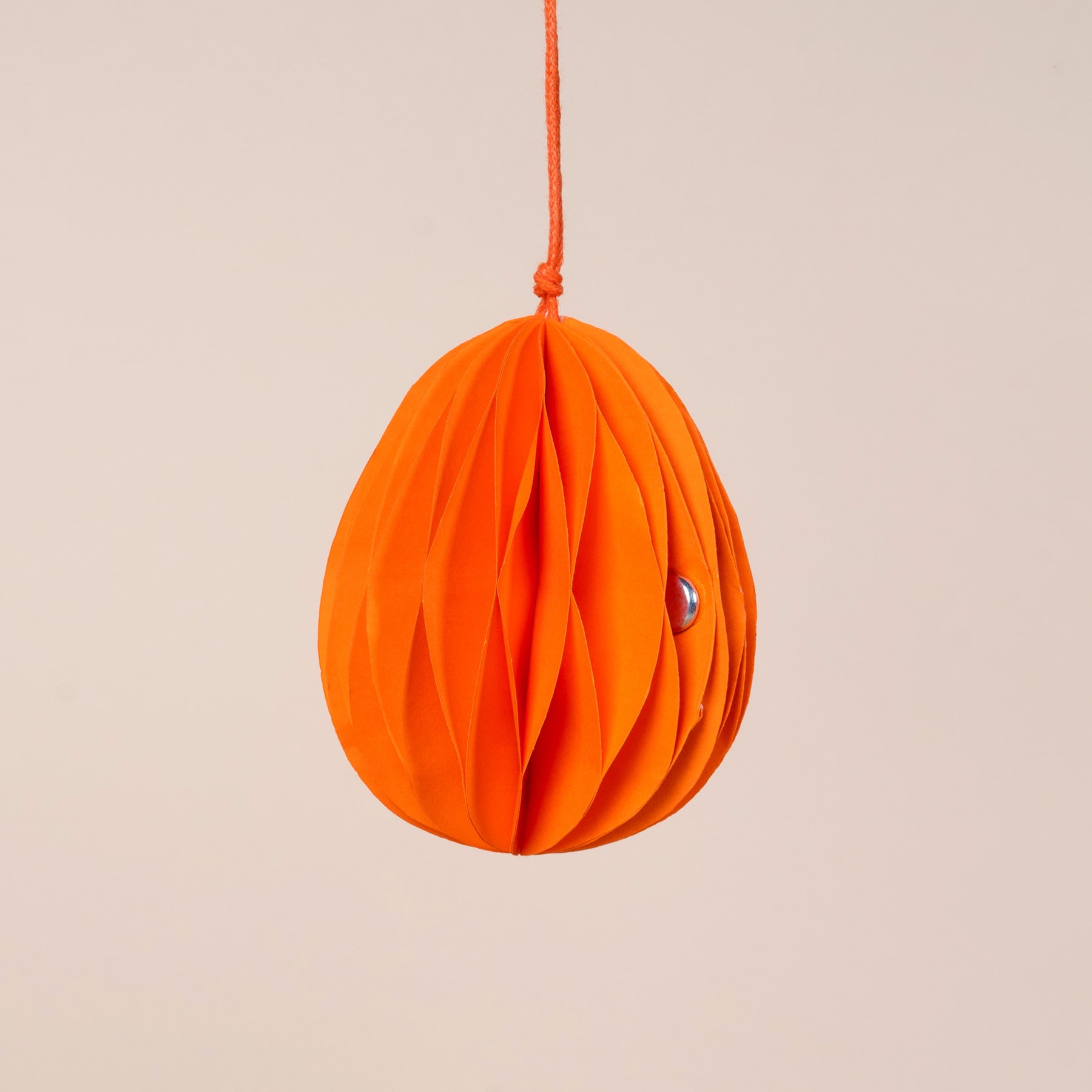 Ein leuchtendes, plissiertes orangefarbenes Ornament hängt vor einem neutralen, beigen Hintergrund.