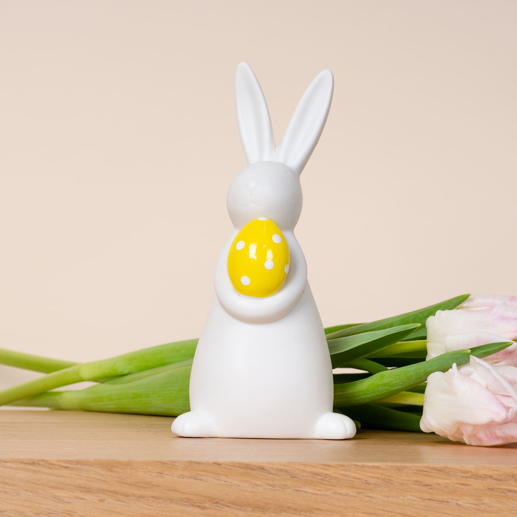 Eine weiße Keramik-Kaninchenfigur, die ein gelbes Osterei hält, umgeben von frischen Tulpen auf einer Holzoberfläche.