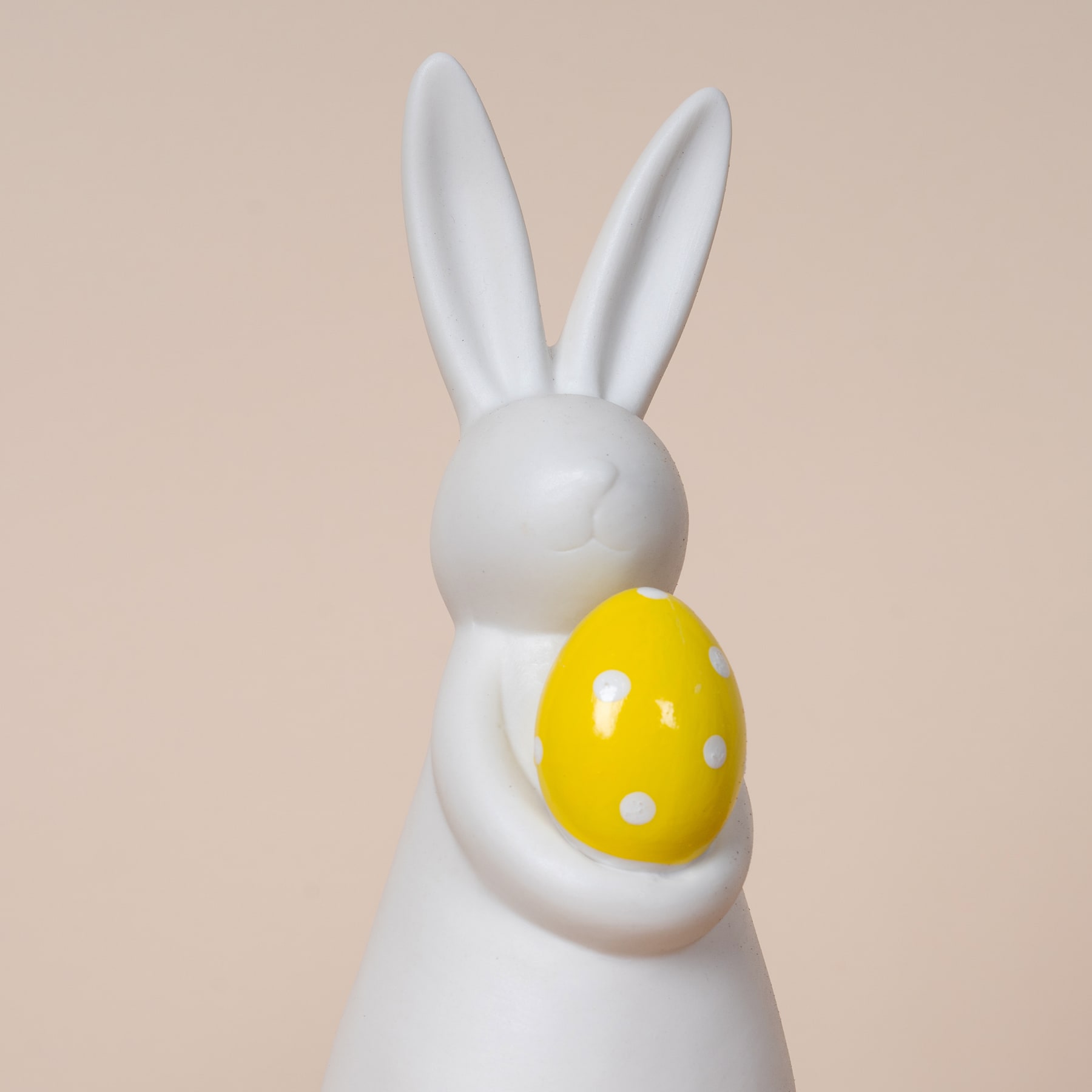 Eine weiße Kaninchenfigur aus Keramik, die vor einem beigen Hintergrund ein gelb gepunktetes Osterei hält.