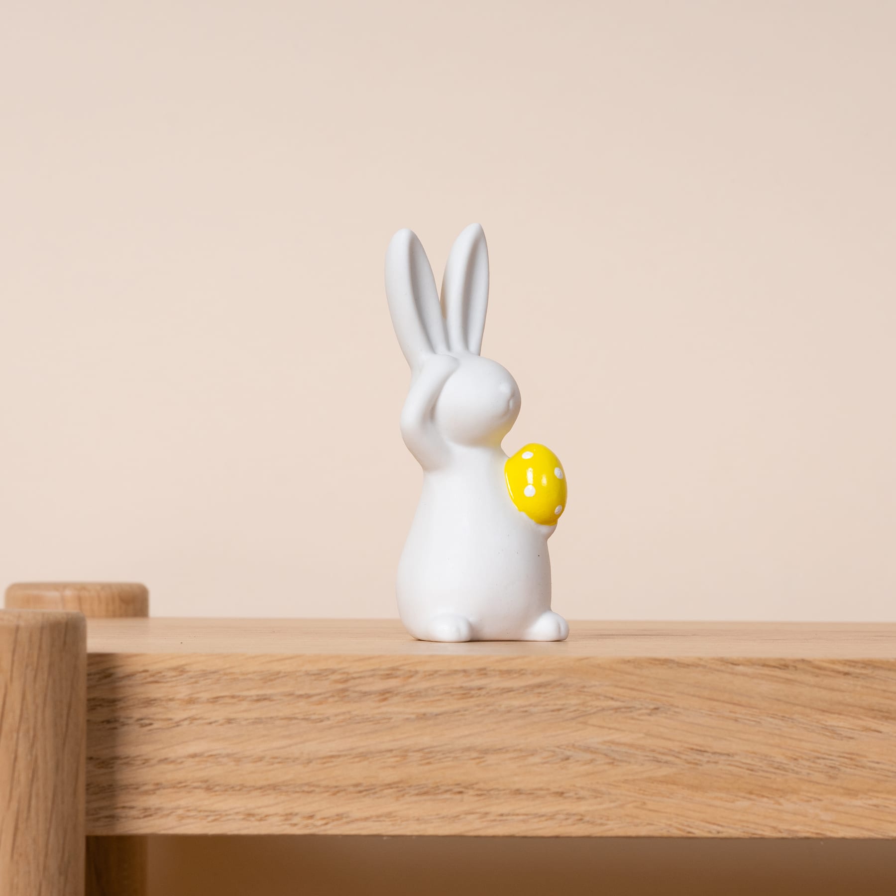 Eine weiße Keramik-Kaninchenfigur, die ein gelbes Ei hält, steht auf einer Holzoberfläche vor einem beigen Hintergrund.