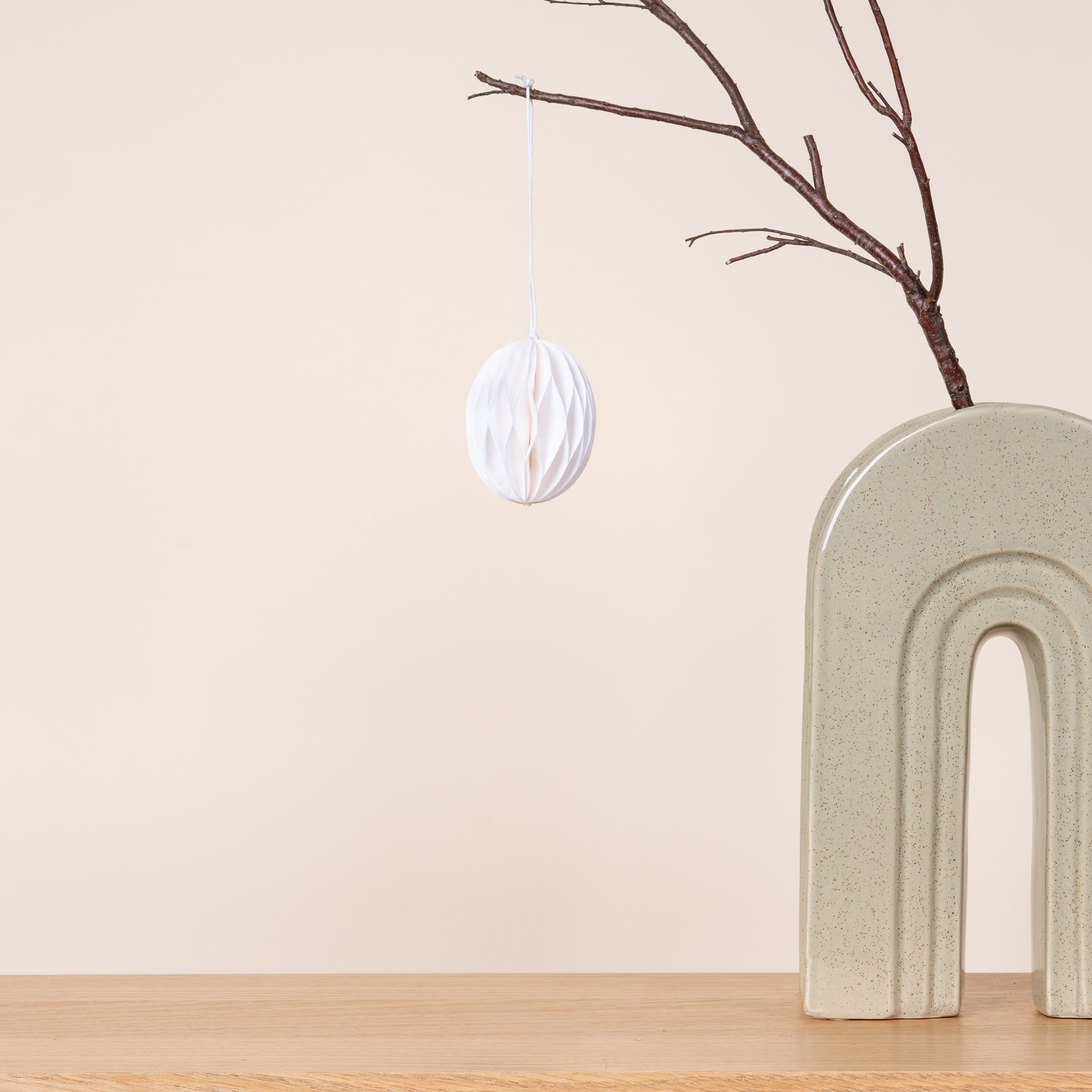 Ein weißes, strukturiertes, eiförmiges Ornament hängt an einem kahlen Ast vor einer Wand in neutralen Tönen, im Vordergrund steht eine minimalistische Keramikskulptur.