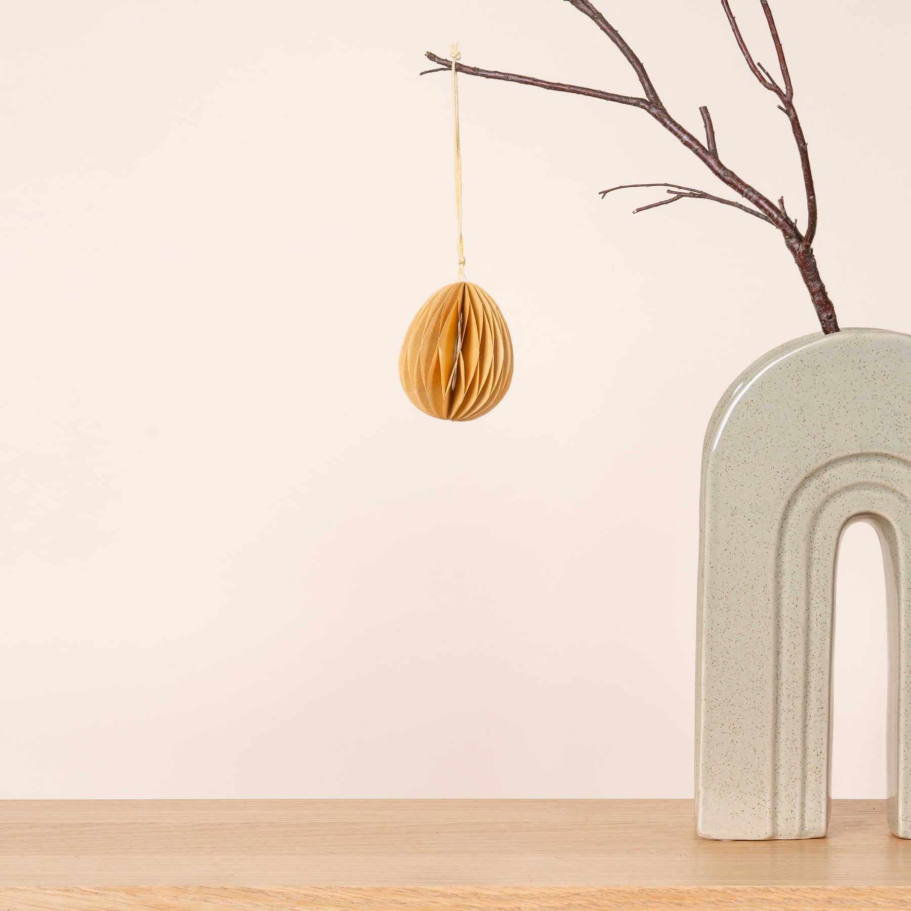 Ein gelbes, wabenförmiges Ornament hängt an einem kahlen Ast vor einer schlichten weißen Wand, im Hintergrund steht eine minimalistische Betonskulptur.