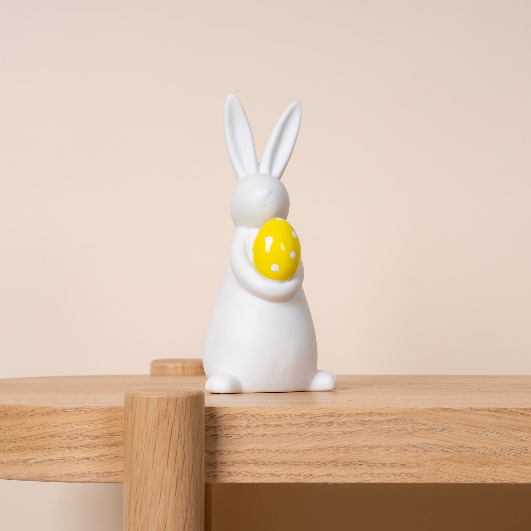 Eine weiße Keramik-Kaninchenfigur mit einem gelben Osterei steht auf einem Holztisch vor einem hellbeigen Hintergrund.