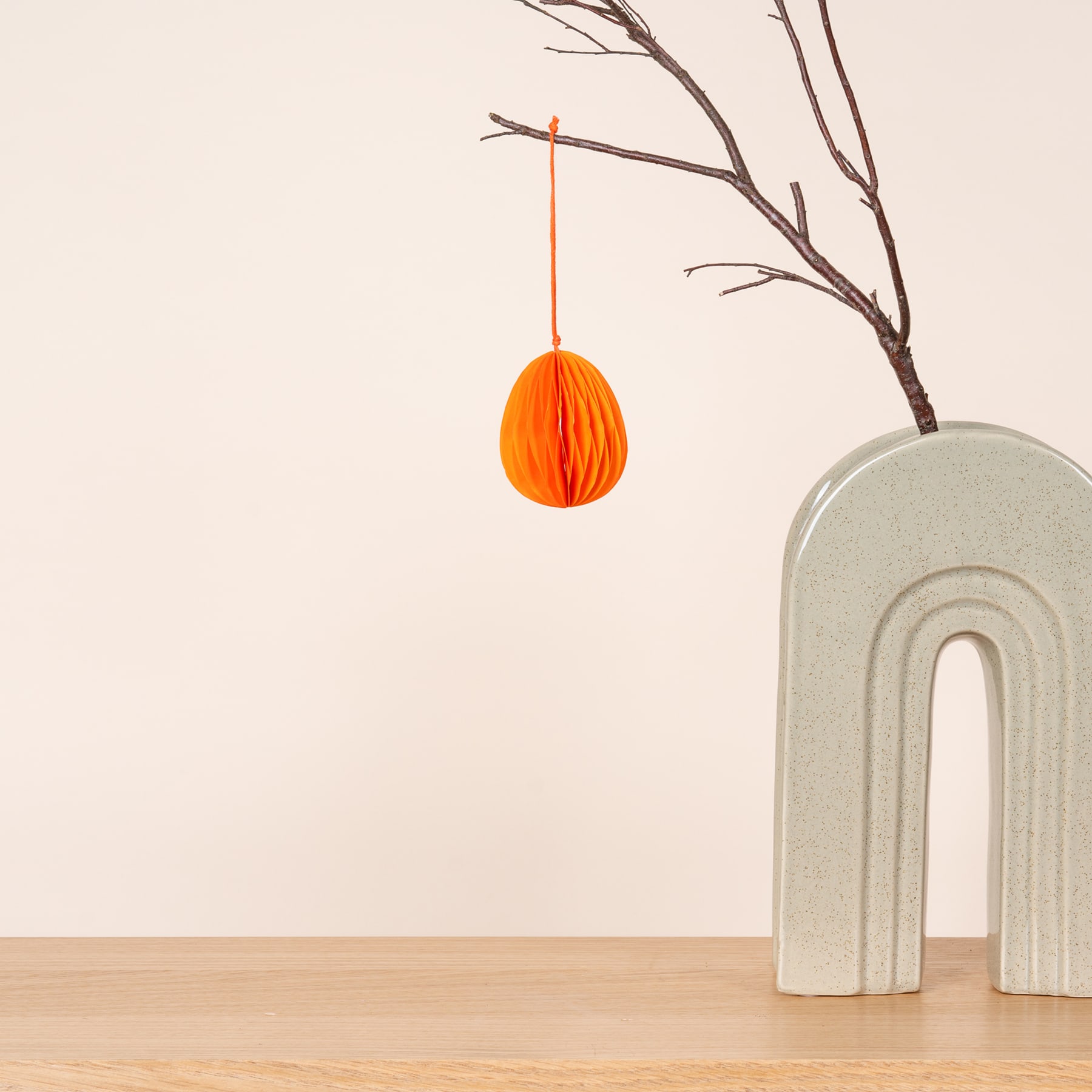 Ein leuchtend orangefarbenes, wabenförmiges Ornament hängt an einem kahlen Ast vor einer schlichten weißen Wand, im Vordergrund steht eine minimalistische Betonskulptur.