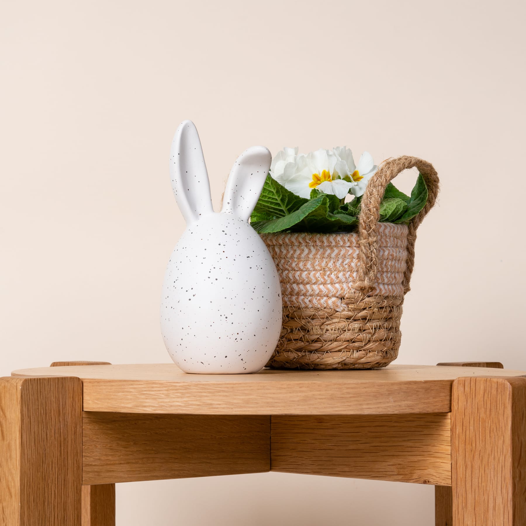 Eine weiße Kaninchenfigur aus Keramik steht auf einem Holzregal neben einem geflochtenen Korb voller grüner Blätter und weißer Blumen.
