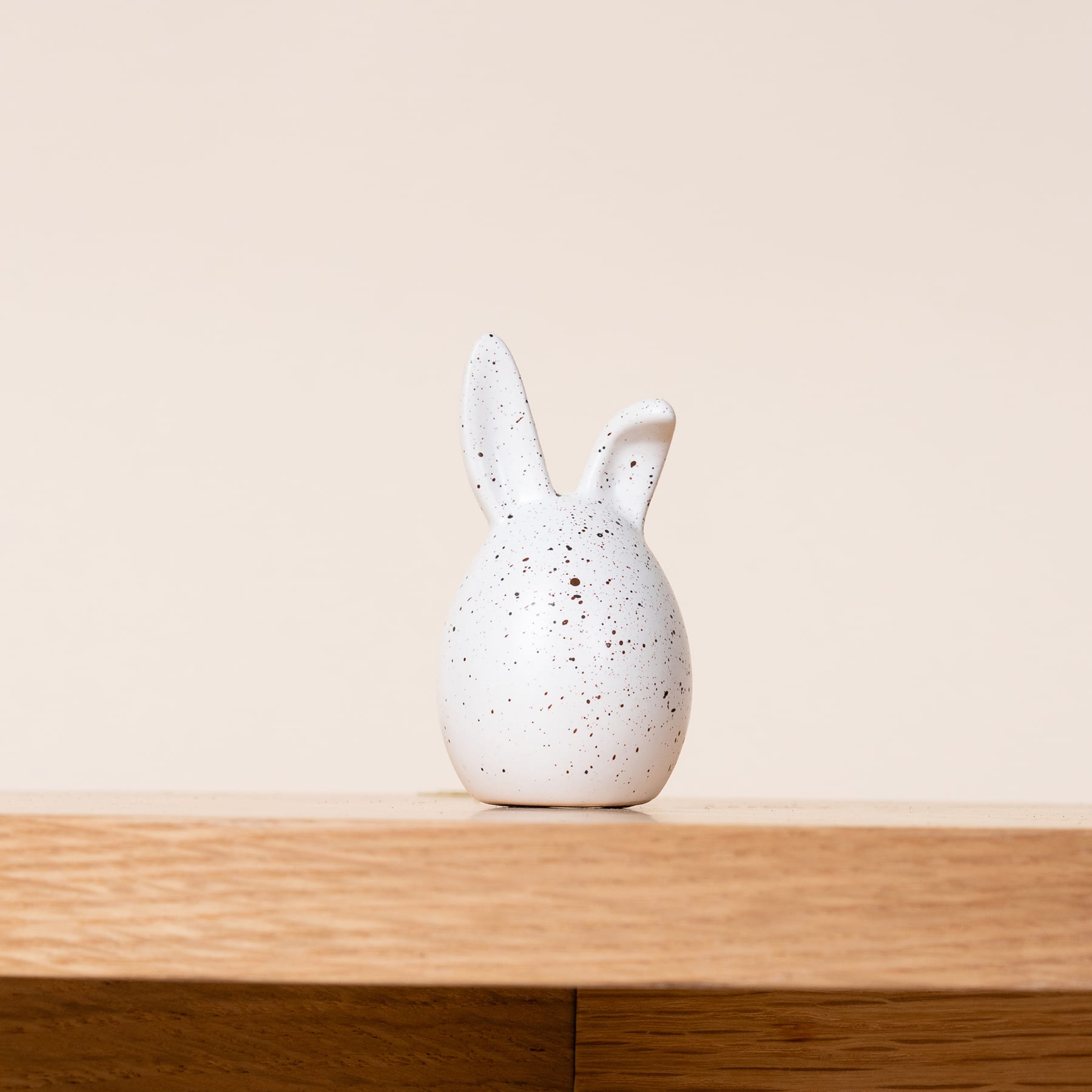 Eine weiße Kaninchenfigur aus Keramik sitzt auf einer Holzoberfläche vor einem schlichten beigen Hintergrund.