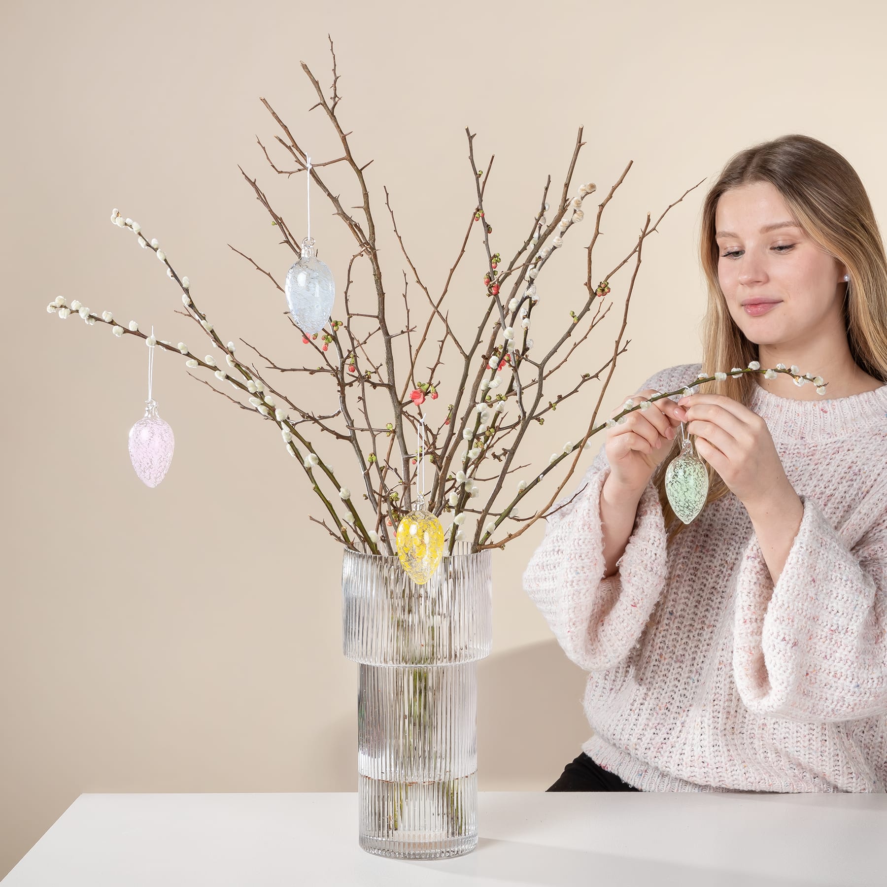 Eine junge Frau in einem kuscheligen Pullover dekoriert vor einem neutralen Hintergrund eine Vase mit kahlen Zweigen, die mit bunten Ostereiern geschmückt sind.