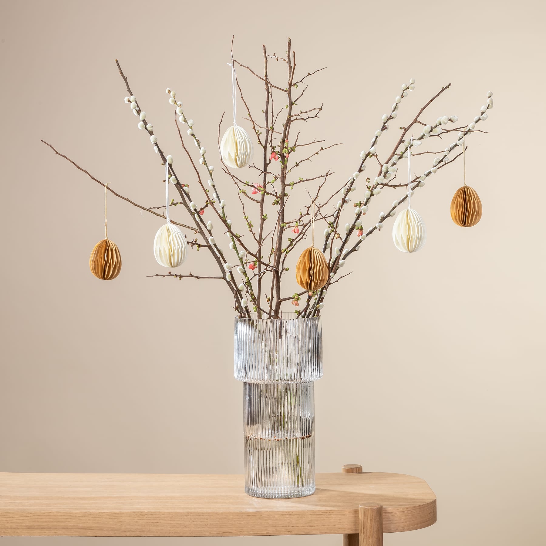 Eine Glasvase mit kahlen Zweigen und hängenden Ornamenten steht auf einem Holztisch vor einem beigen Hintergrund.