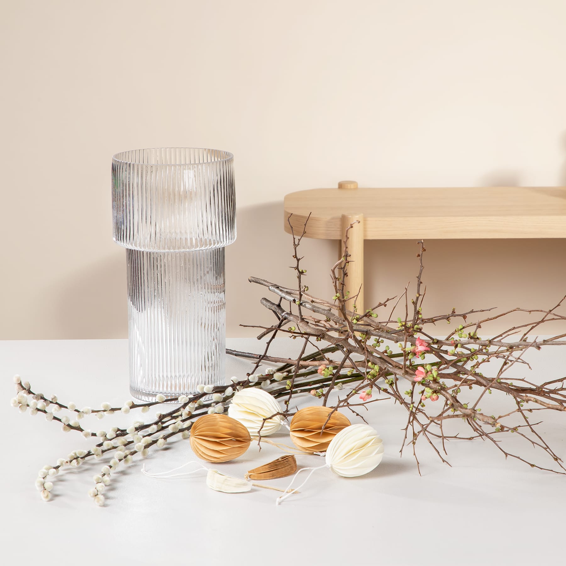 Auf einem Holzregal steht eine Glasvase mit Rippenmuster, daneben ein Zweig mit Frühlingsblüten und mehrere Macarons, die auf der Oberfläche angeordnet sind.