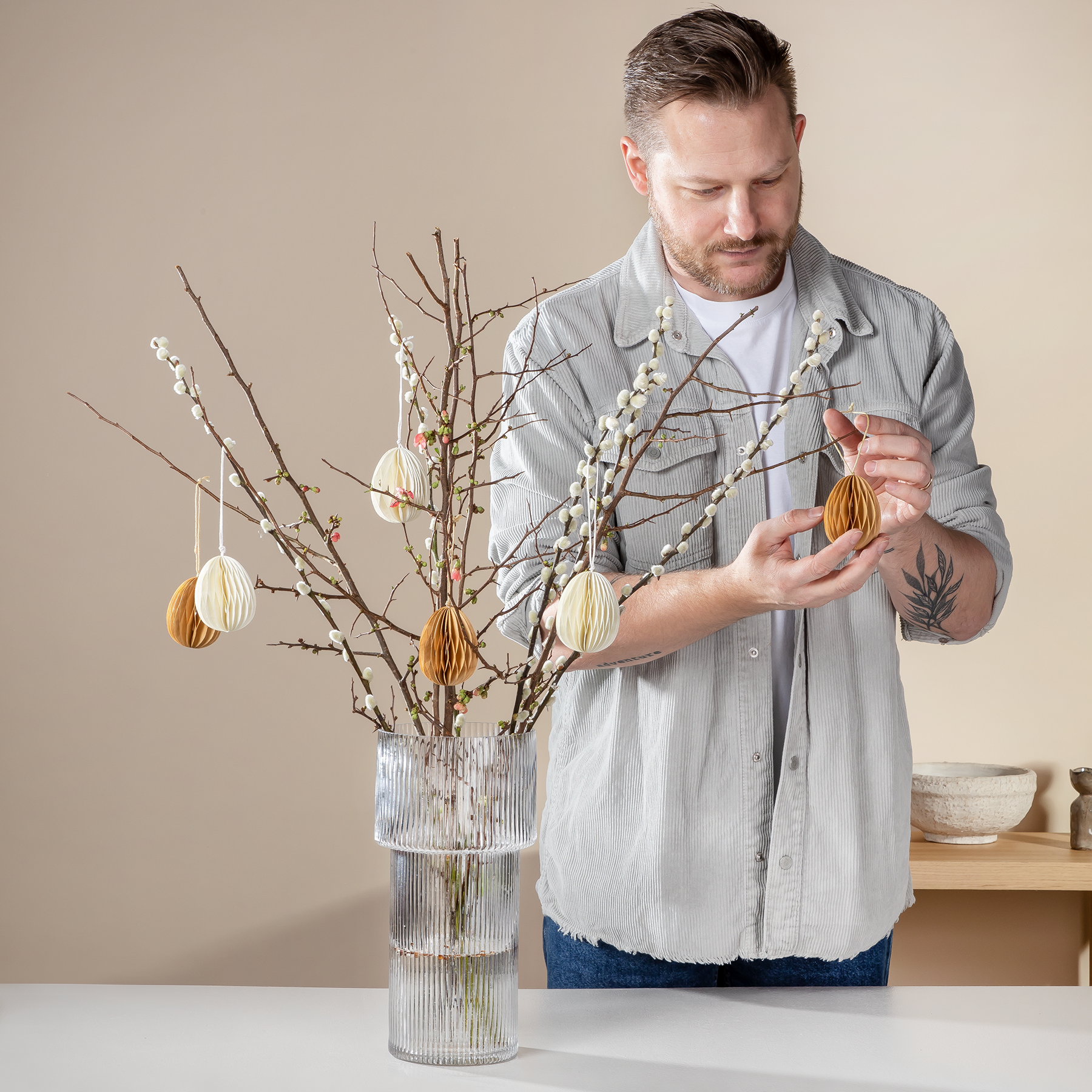 Ein Mann mit Bart und Tattoos arrangiert dekorative Zweige mit Ostereiern in einer Vase auf einem Tisch vor einem schlichten beigen Hintergrund.