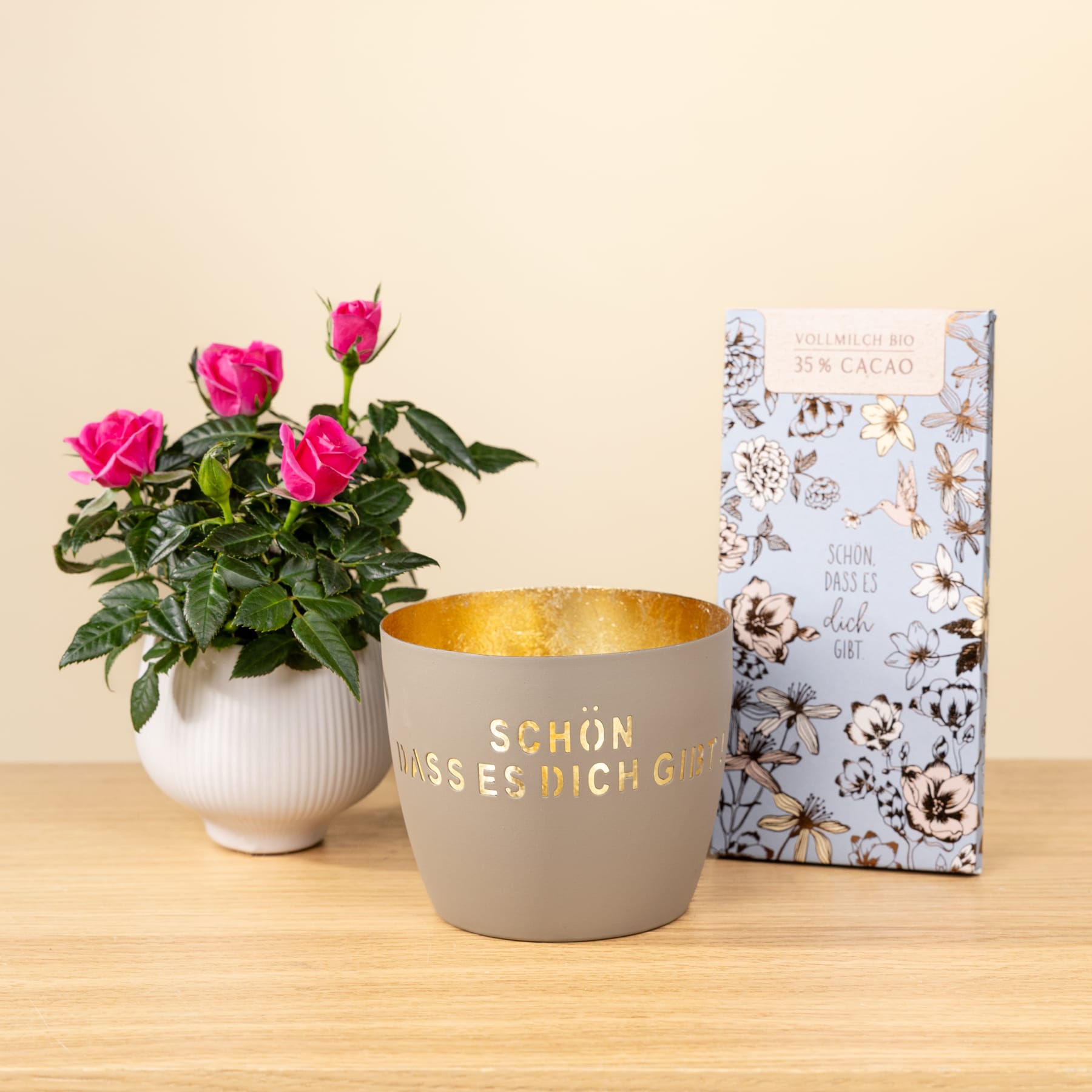 Das Bild zeigt eine Topfpflanze mit leuchtend rosa Rosen, einen grau-goldenen Übertopf mit der Aufschrift „Schon dass es dich gibt“ und ein Buch mit Blumenmuster auf einem Holztisch.