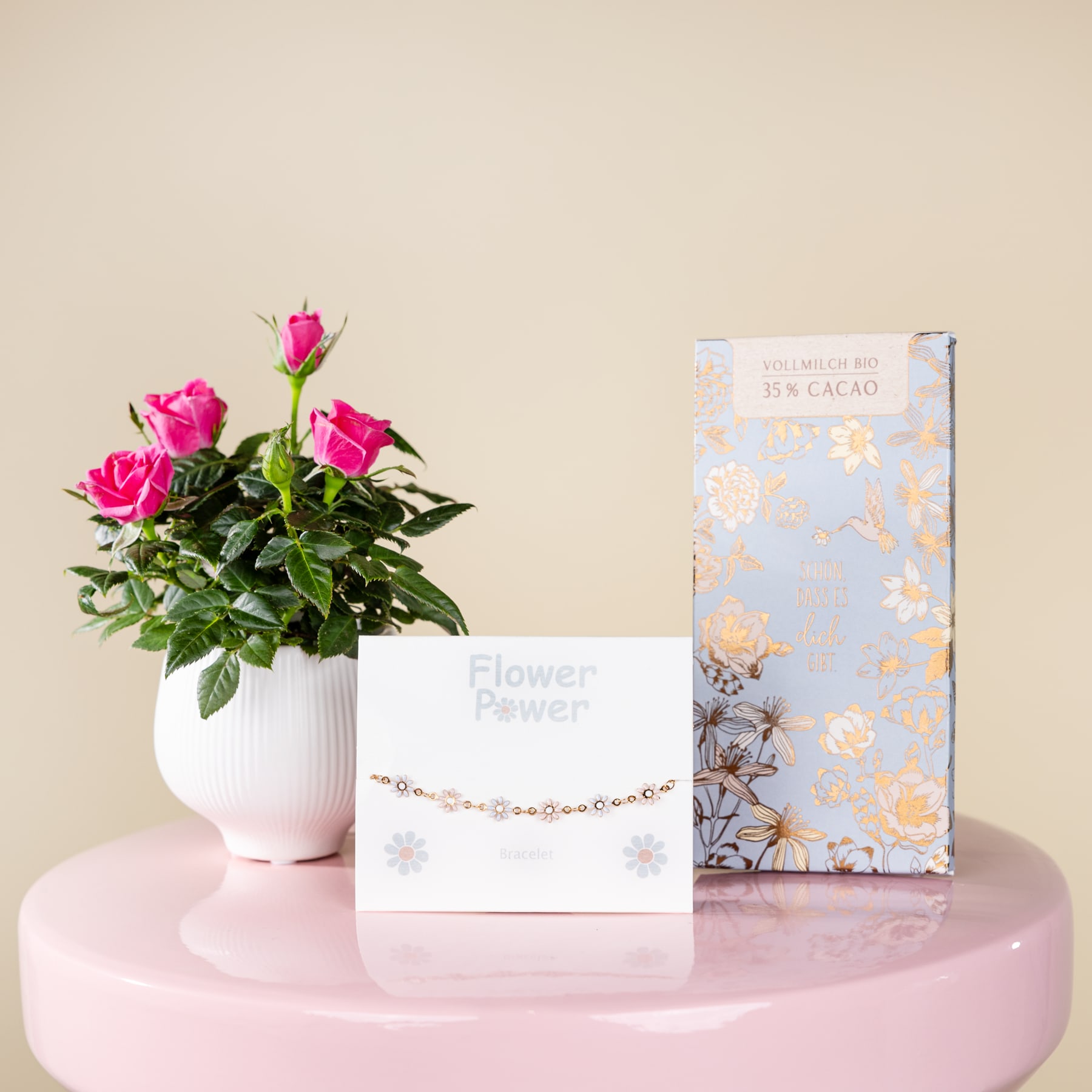 Eine Vase mit leuchtend rosa Rosen steht auf einem rosa Sockel, dazu gibt es eine Grußkarte und eine dekorative Geschenkbox mit Blumenmuster.