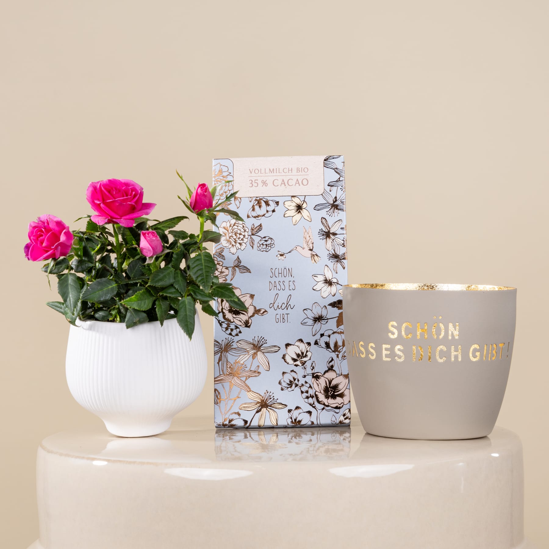Auf einem Tisch steht eine Vase mit leuchtend rosa Rosen, dazu ein Buch und ein dekorativer Keramiktopf, alles vor einem neutralen beigen Hintergrund.