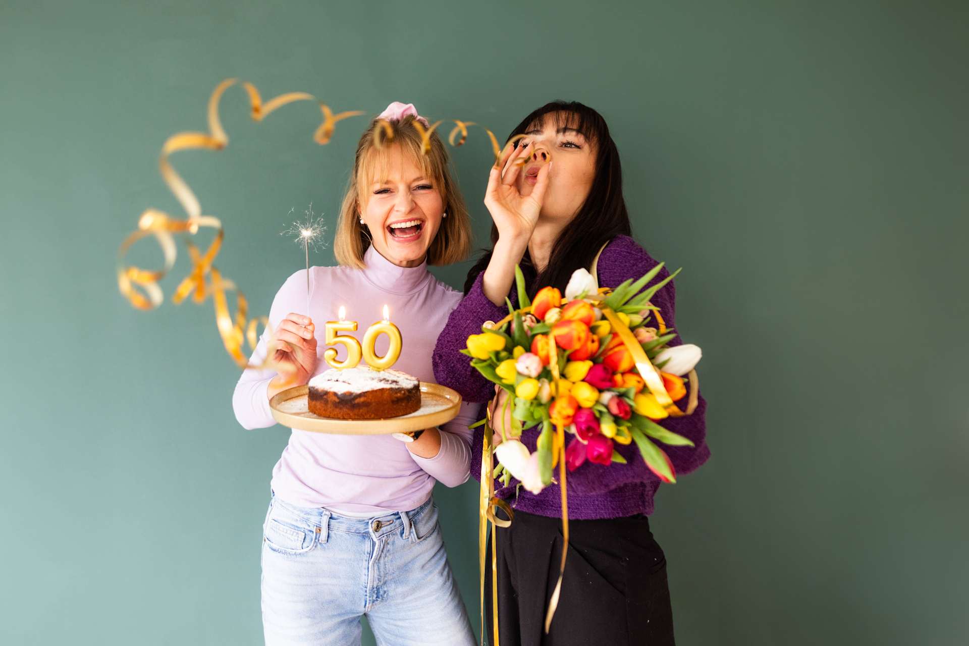 Zwei Frauen, eine hält eine Geburtstagstorte mit einer „50“-Kerze und die andere einen Blumenstrauß, lächeln und feiern gemeinsam vor einem grünen Hintergrund.