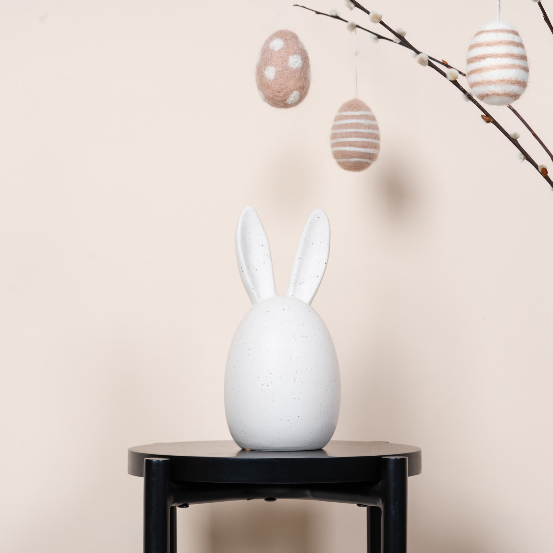Eine weiße Kaninchenfigur aus Keramik sitzt auf einem schwarzen Holzständer, im Hintergrund hängen dekorative Ostereier von Zweigen.