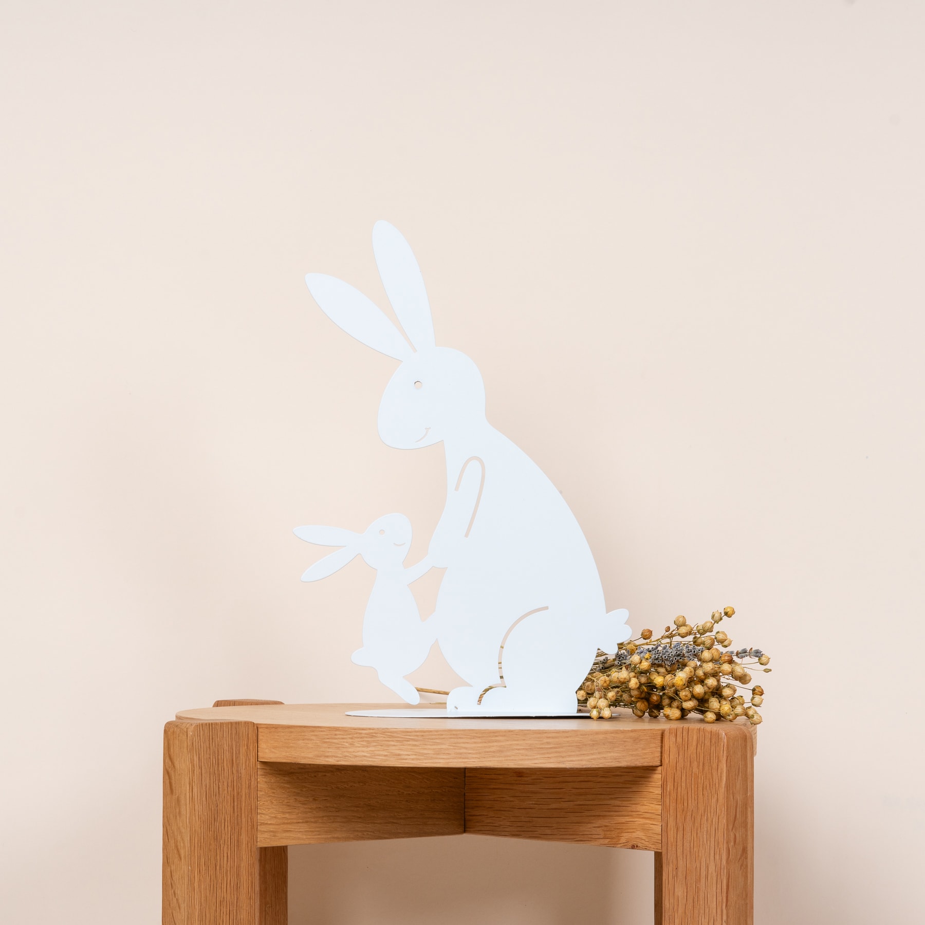 Vor einem hellbeigen Hintergrund steht ein Holztisch mit einer weißen Hasensilhouette und einem Strauß getrockneter Blumen.