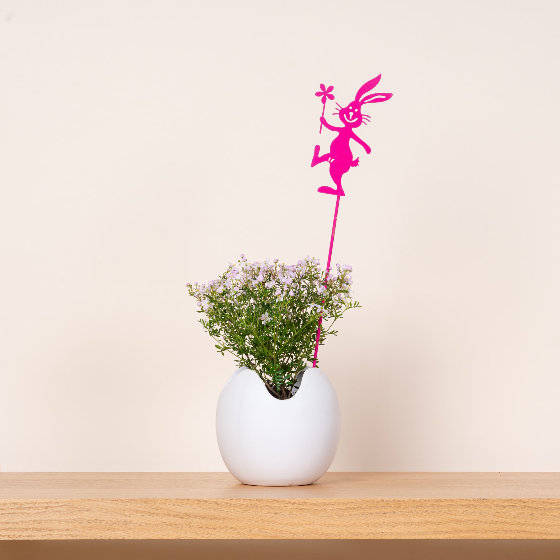 Eine weiße Keramikvase auf einem Holztisch enthält einen Strauß kleiner weißer Blumen. Auf dem Arrangement steht die rosa Metallskulptur einer kaninchenähnlichen Figur.