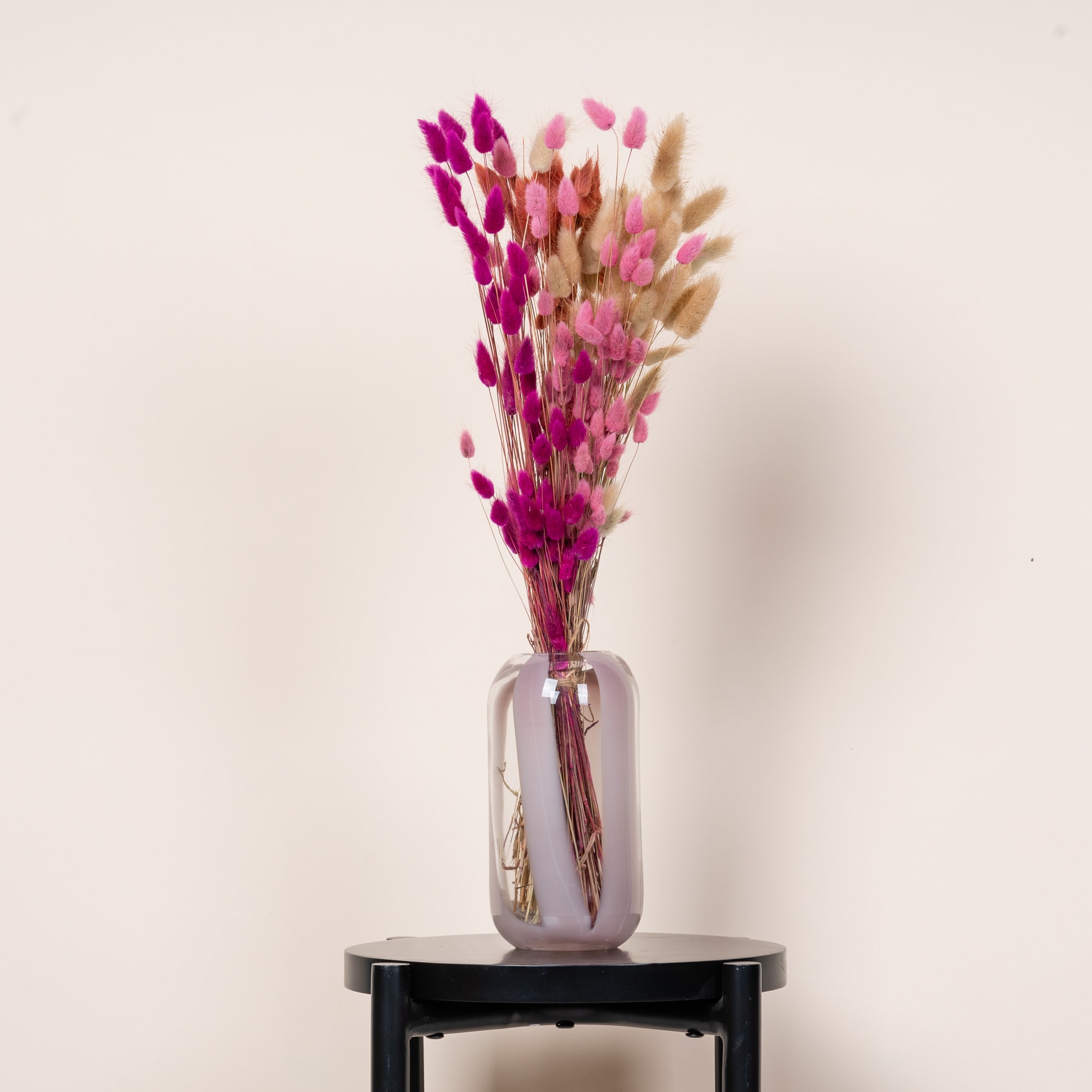 Eine Vase mit leuchtend rosa und beigen Trockenblumen steht auf einer dunklen Holzoberfläche vor einem schlichten weißen Hintergrund.