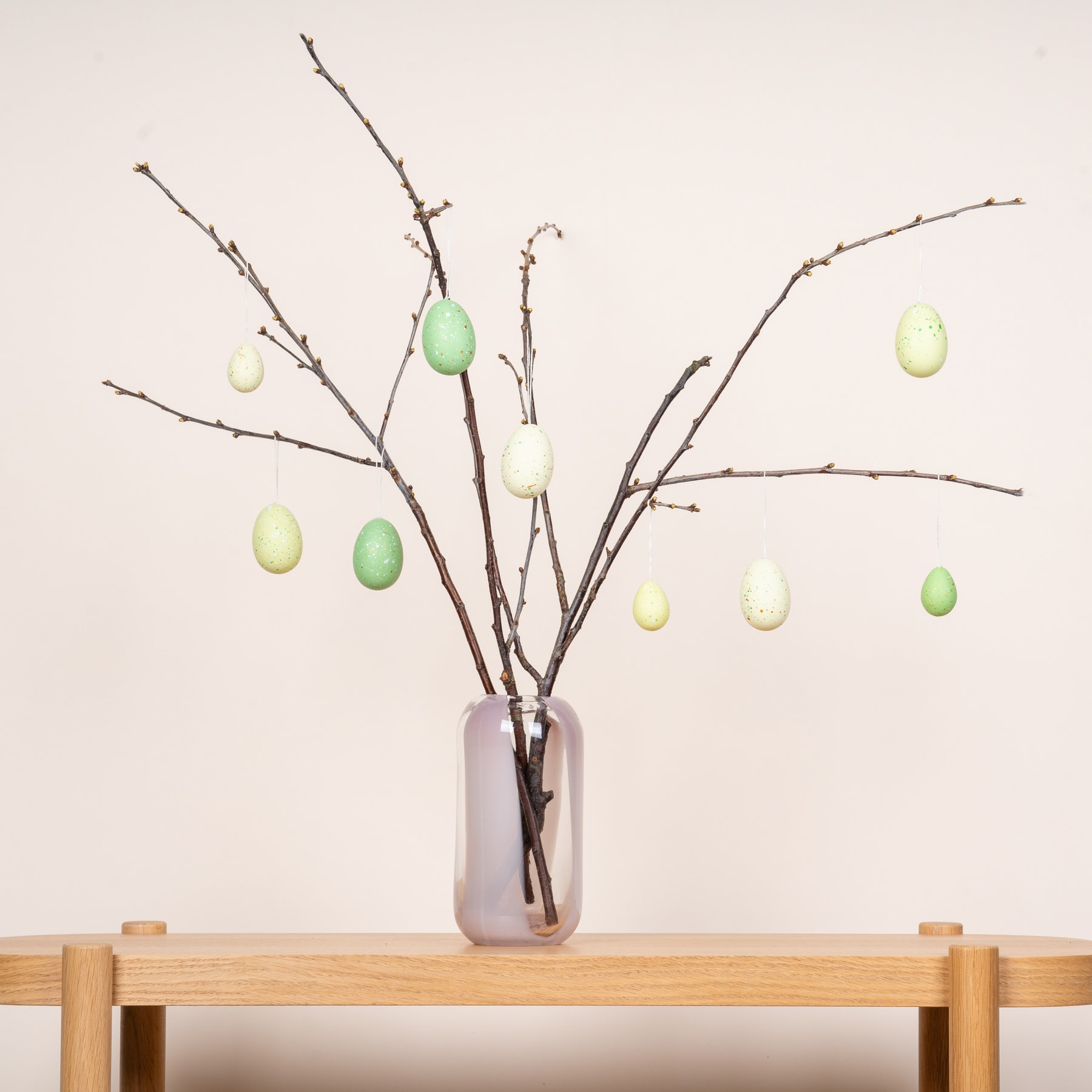 Eine Vase mit kahlen Zweigen, die mit bunten Ostereiern geschmückt sind, steht auf einem Holztisch vor einem schlichten Hintergrund.