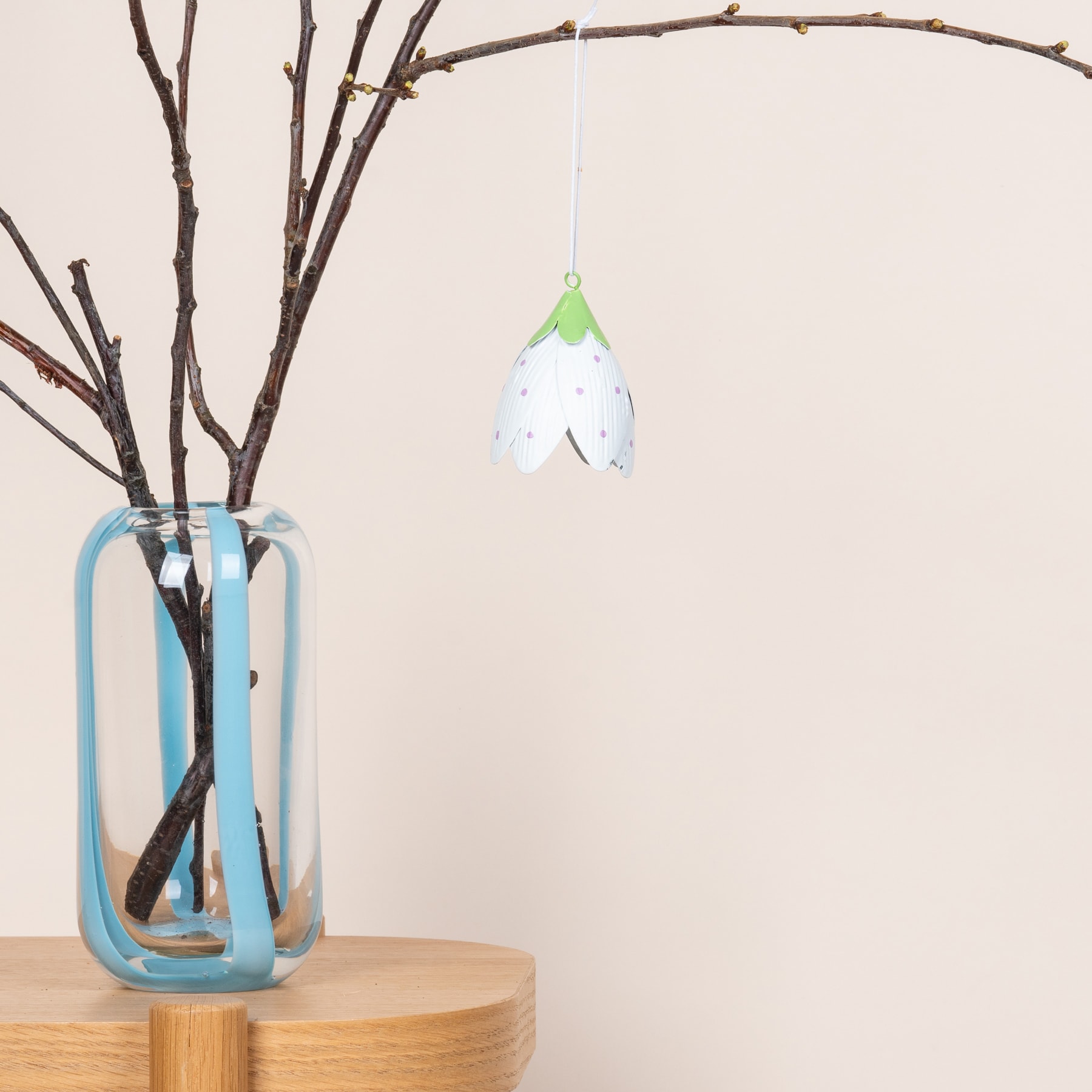 Auf einer Holzoberfläche steht eine Glasvase mit kahlen Zweigen, an den Zweigen hängt ein hängendes Ornament in Form eines Blattes.