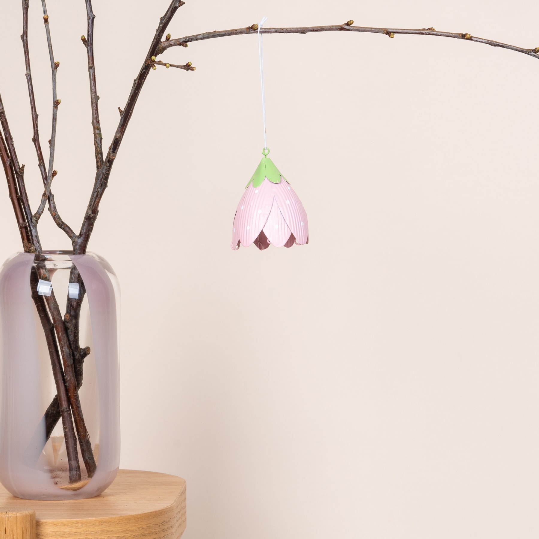 Ein zartes rosa und grünes, blumenförmiges Ornament hängt an einem Zweig in einer Glasvase vor einem schlichten Hintergrund.