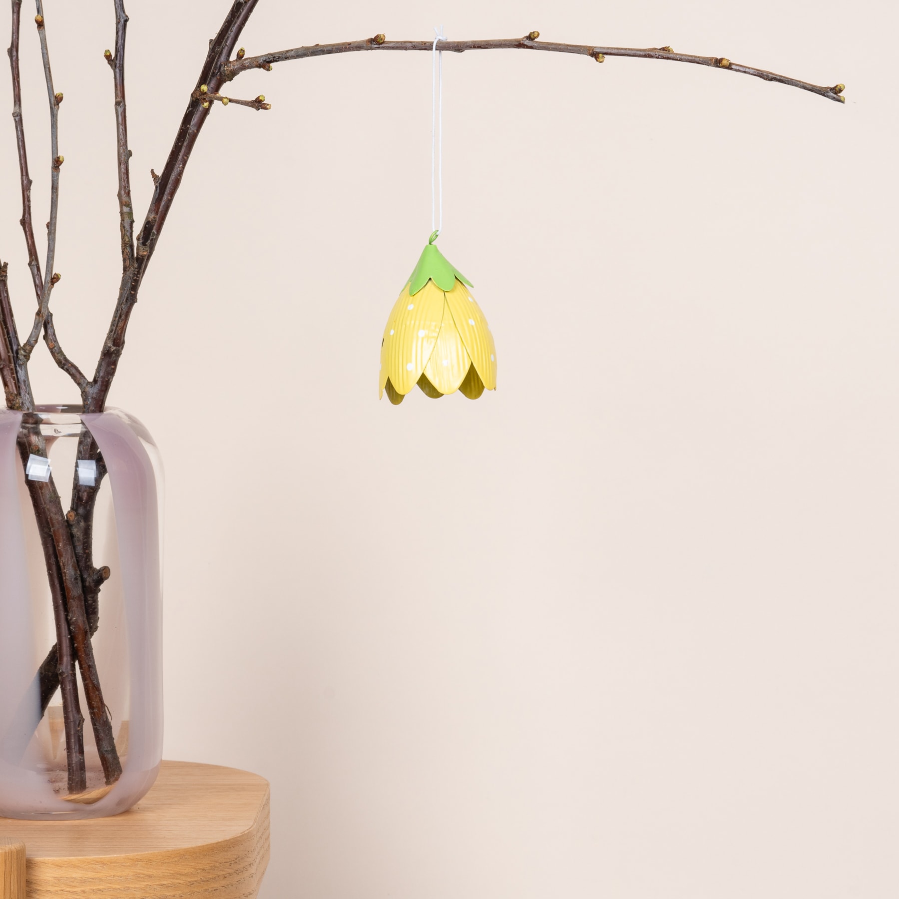 Ein hängendes gelbes, blumenförmiges Ornament, das an einem Zweig in einer Vase auf einer Holzoberfläche vor einem schlichten weißen Hintergrund hängt.