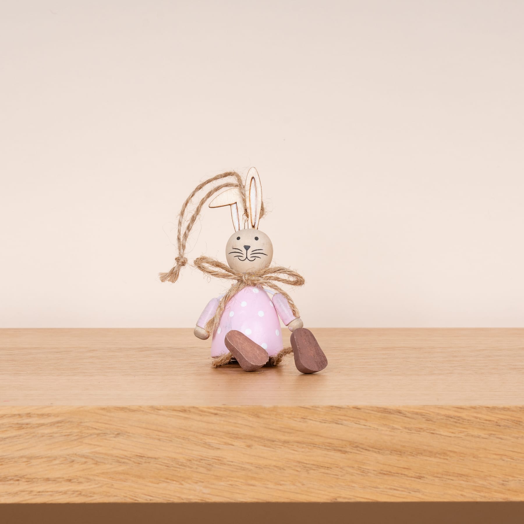 Ein Plüschkaninchen mit langen Ohren und einem rosa Outfit sitzt auf einer Holzoberfläche vor einem schlichten beigen Hintergrund.