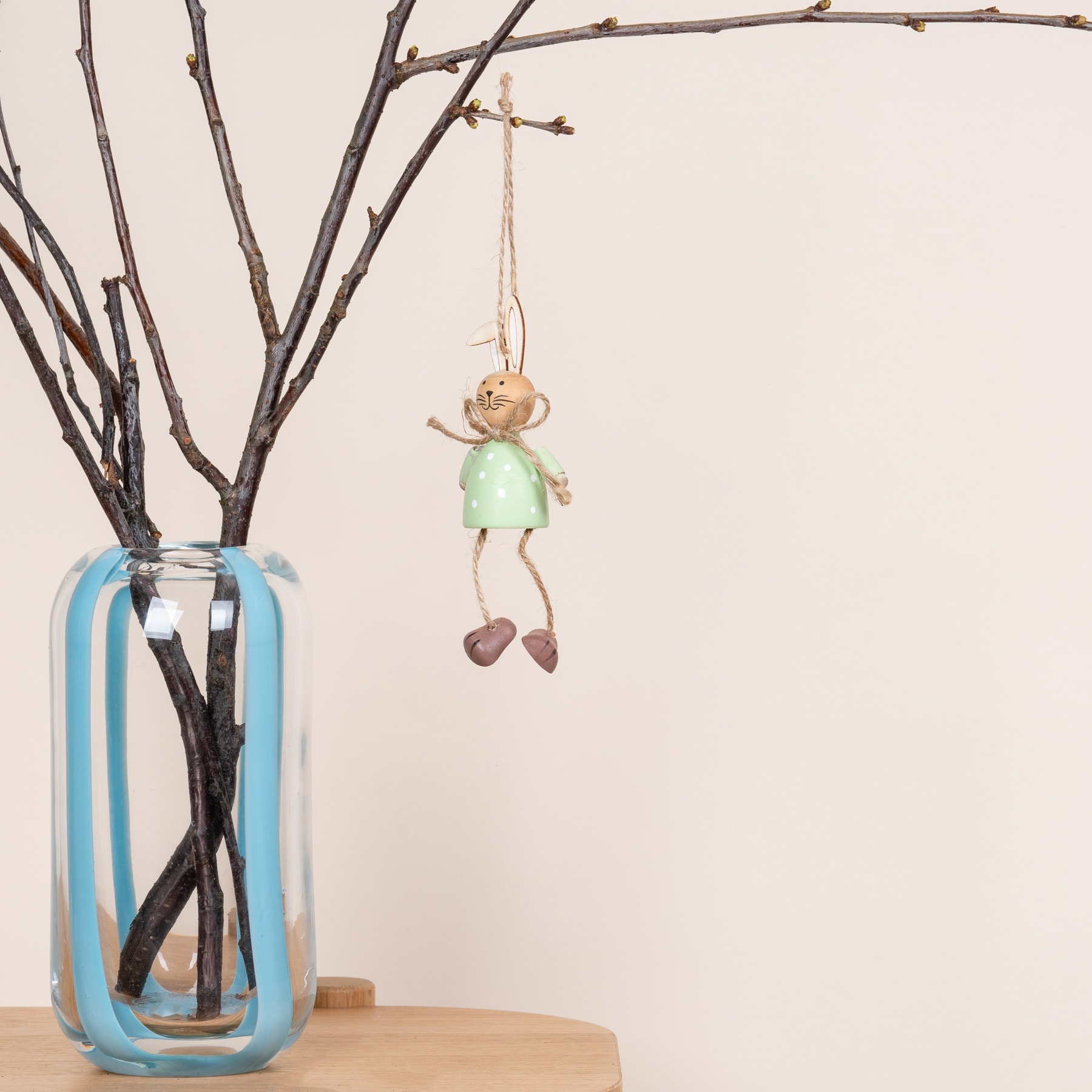Eine kleine, skurrile Puppenfigur hängt an einem Ast in einer Vase voller kahler Zweige vor einem schlichten weißen Hintergrund.