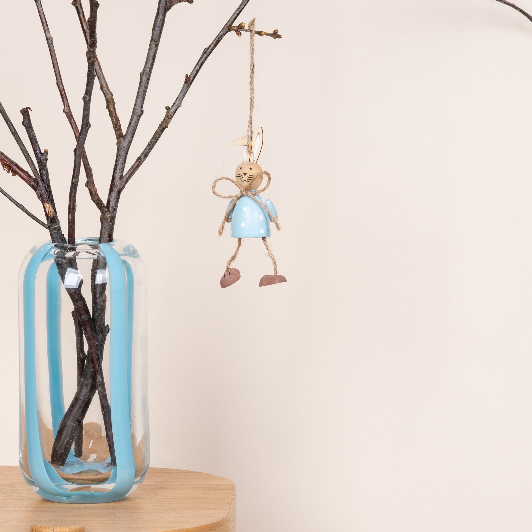 Eine blaue Glasvase mit kahlen Zweigen und einem hängenden Ornament in Form einer kleinen Figur in blauem Gewand.