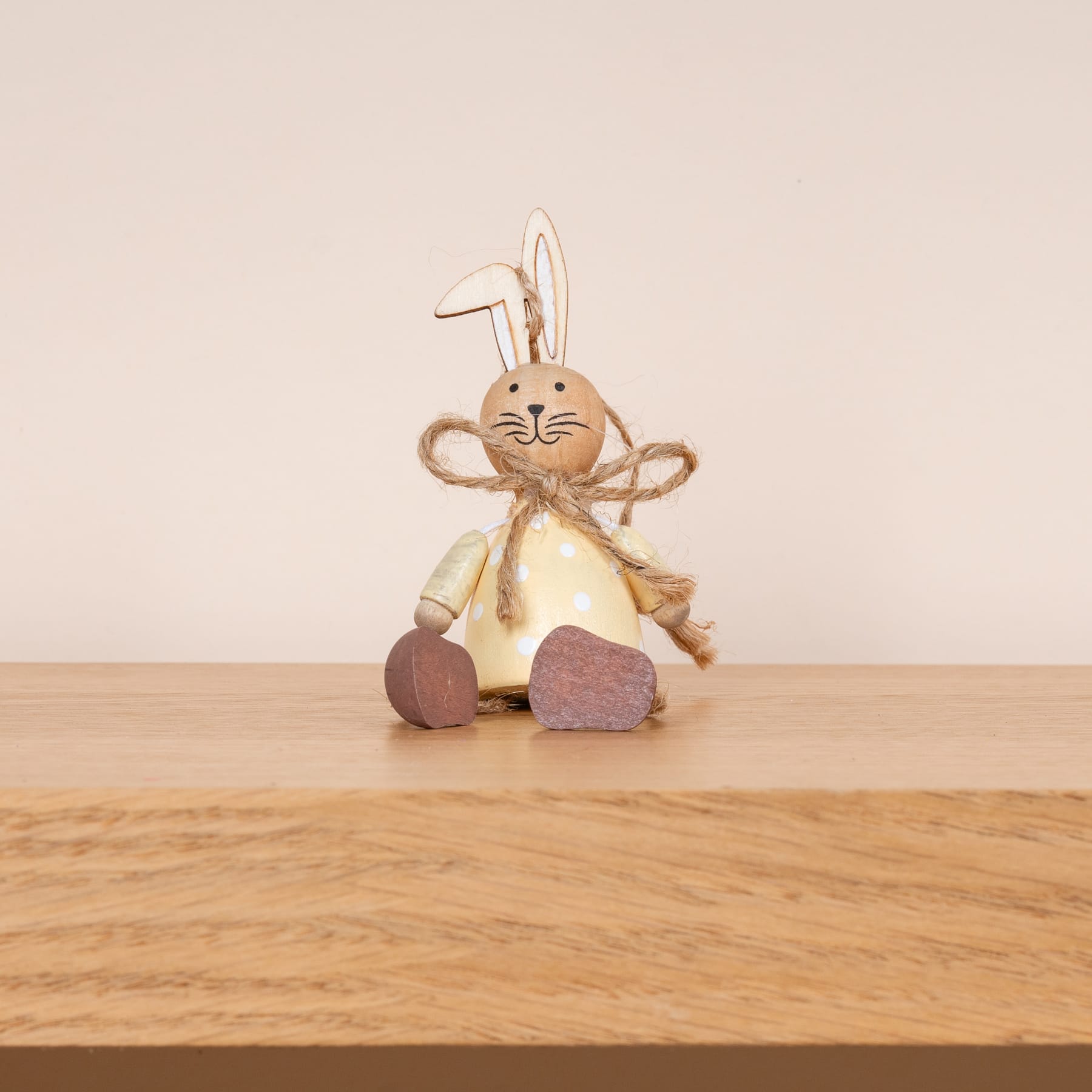 Eine Plüsch-Hasenfigur mit langen Ohren und gelbem Kleid sitzt auf einer Holzoberfläche vor einem beigen Hintergrund.