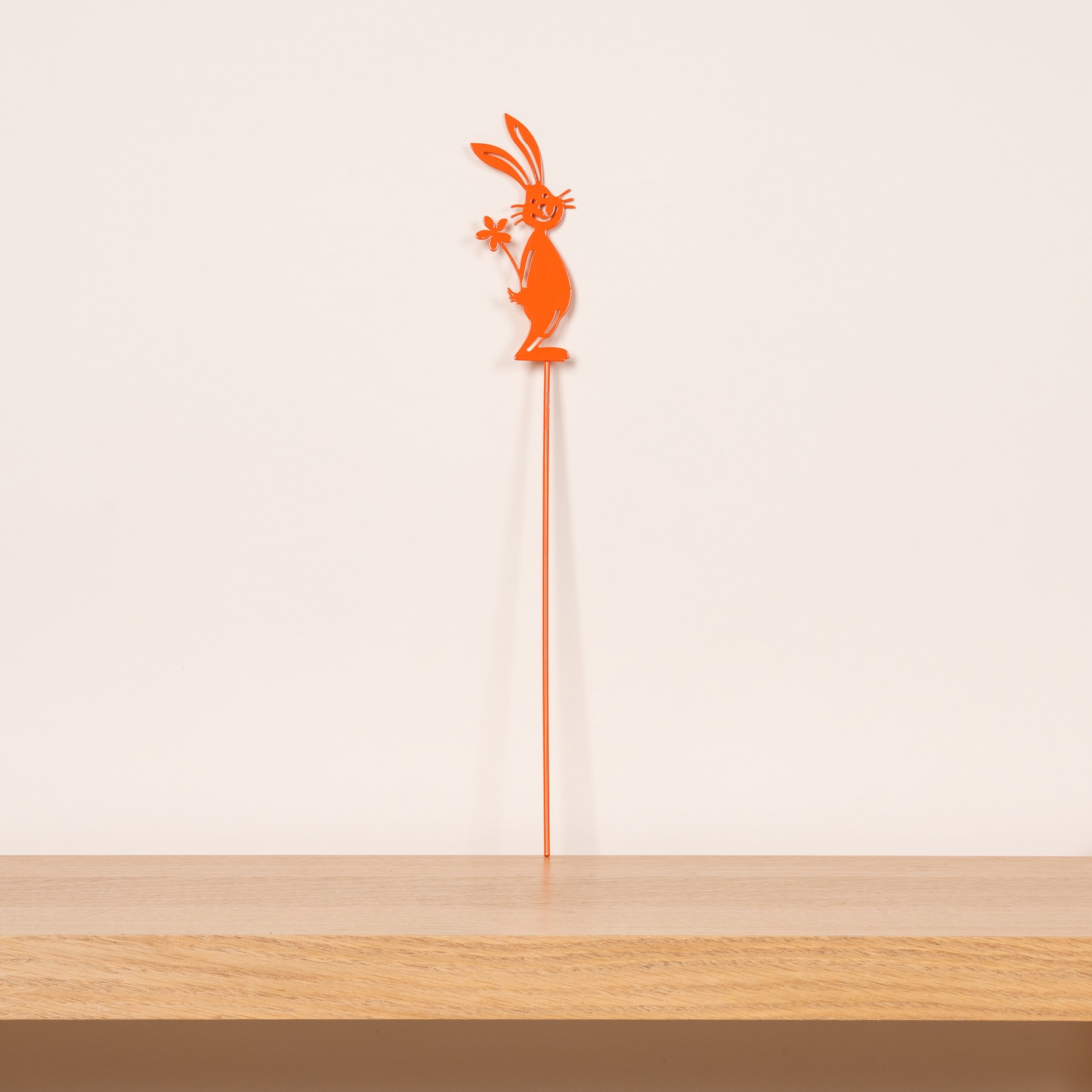 Ein leuchtend orangefarbenes Ornament in Hasenform steht auf einer Holzoberfläche vor einem schlichten, hellen Hintergrund.