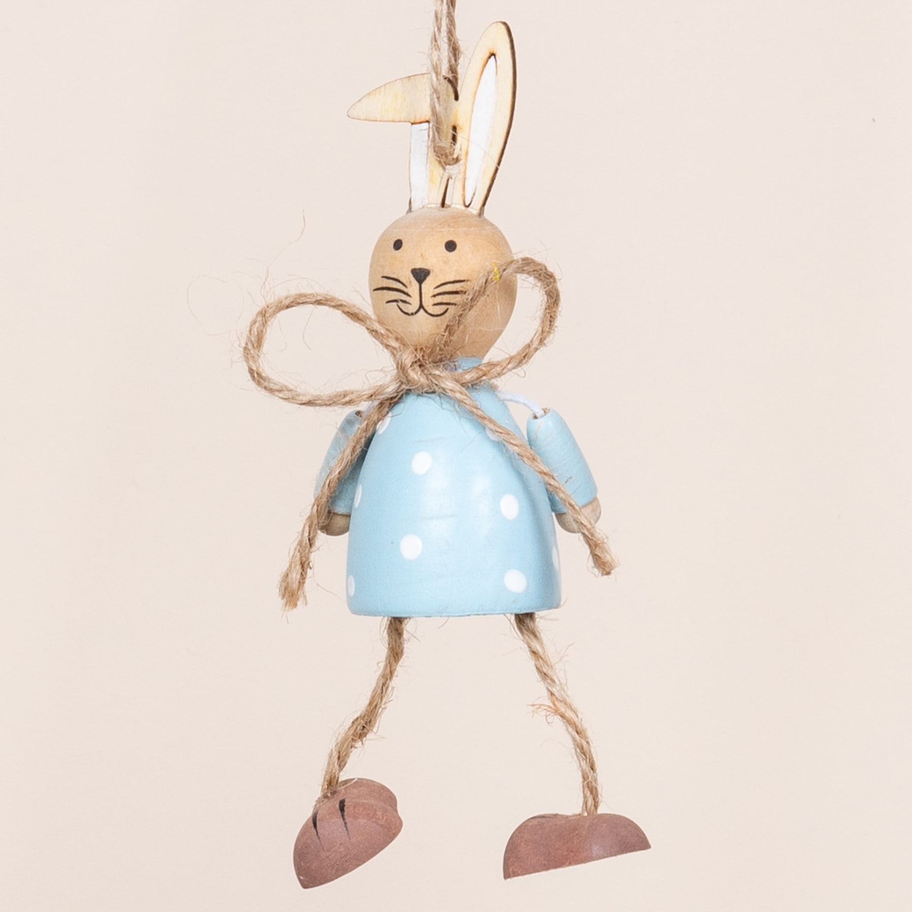 Eine skurrile Kaninchenfigur aus Holz mit blauem Kleid und Seilgliedern hängt vor einem schlichten beigen Hintergrund.