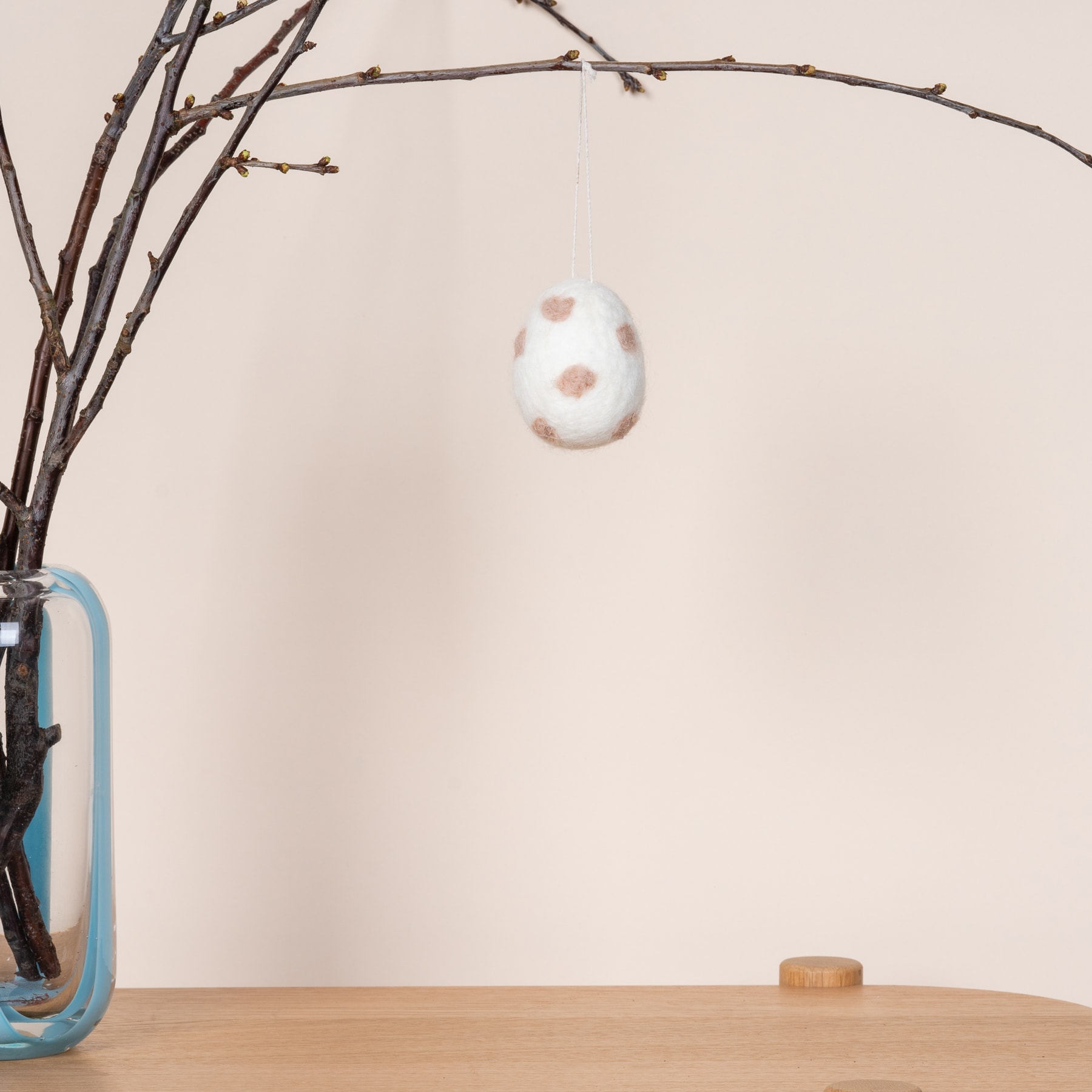 Ein weißes, gesprenkeltes, eiförmiges Ornament hängt an einem Bündel kahler Zweige vor einer schlichten, hellen Wand. Im Hintergrund sind eine Glasvase und ein Holzgegenstand zu sehen.