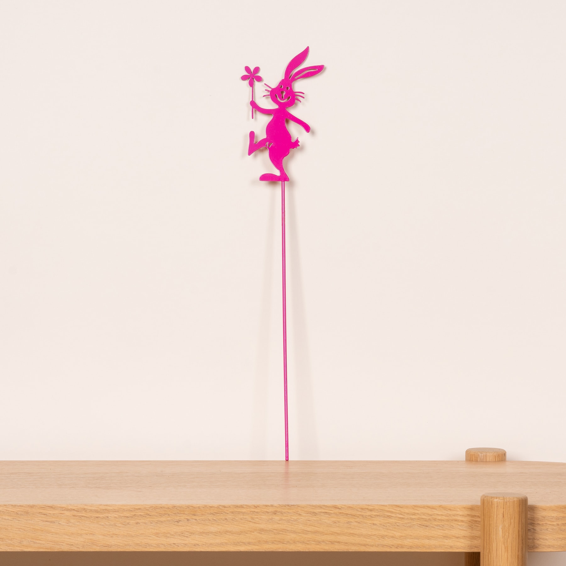 Eine leuchtend rosa, märchenhafte Figur steht auf einer Holzoberfläche vor einem schlichten Hintergrund.
