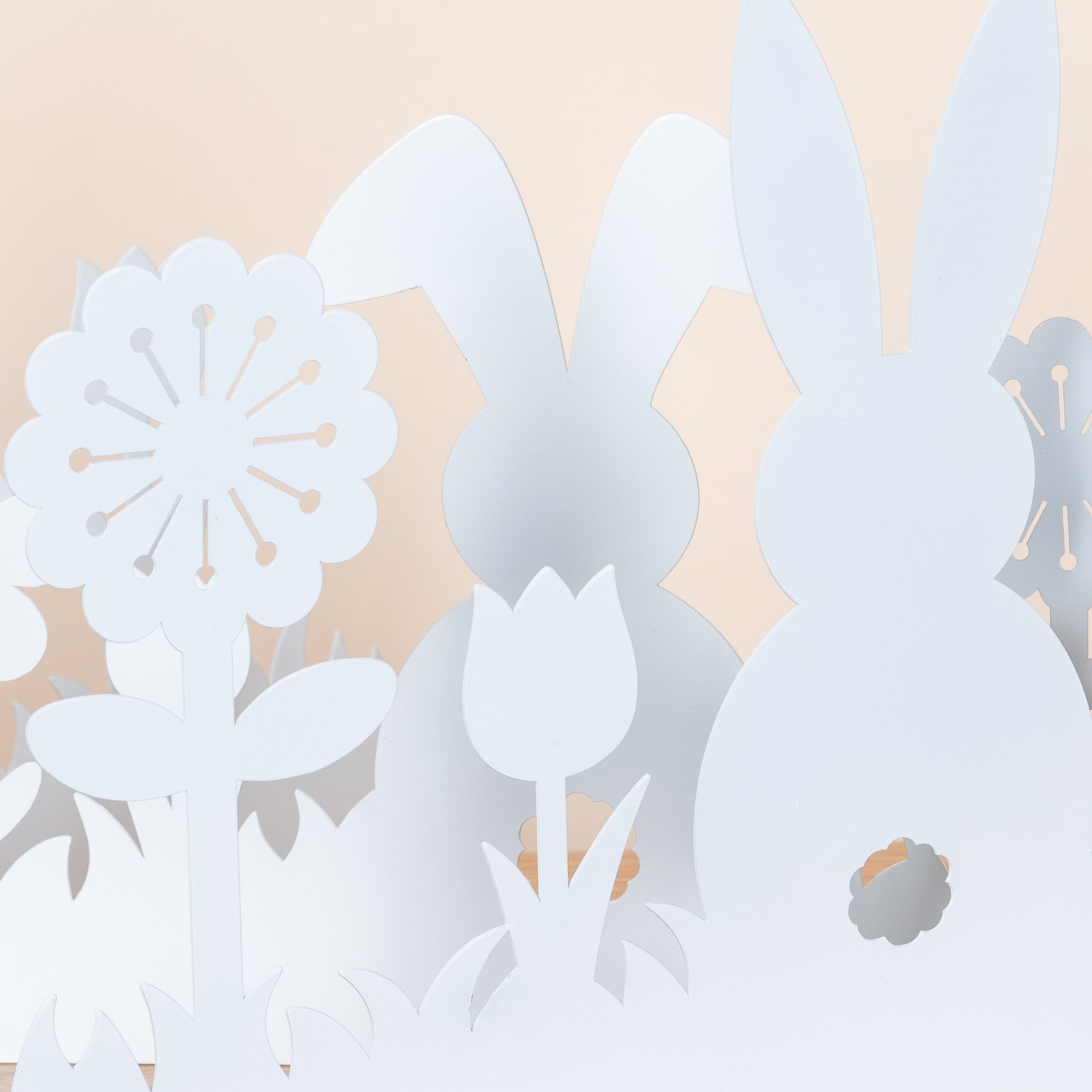 Das Bild zeigt zarte weiße Papierausschnitte verschiedener floraler Formen, darunter Blumen und Blätter, vor einem weichen, pfirsichfarbenen Hintergrund.