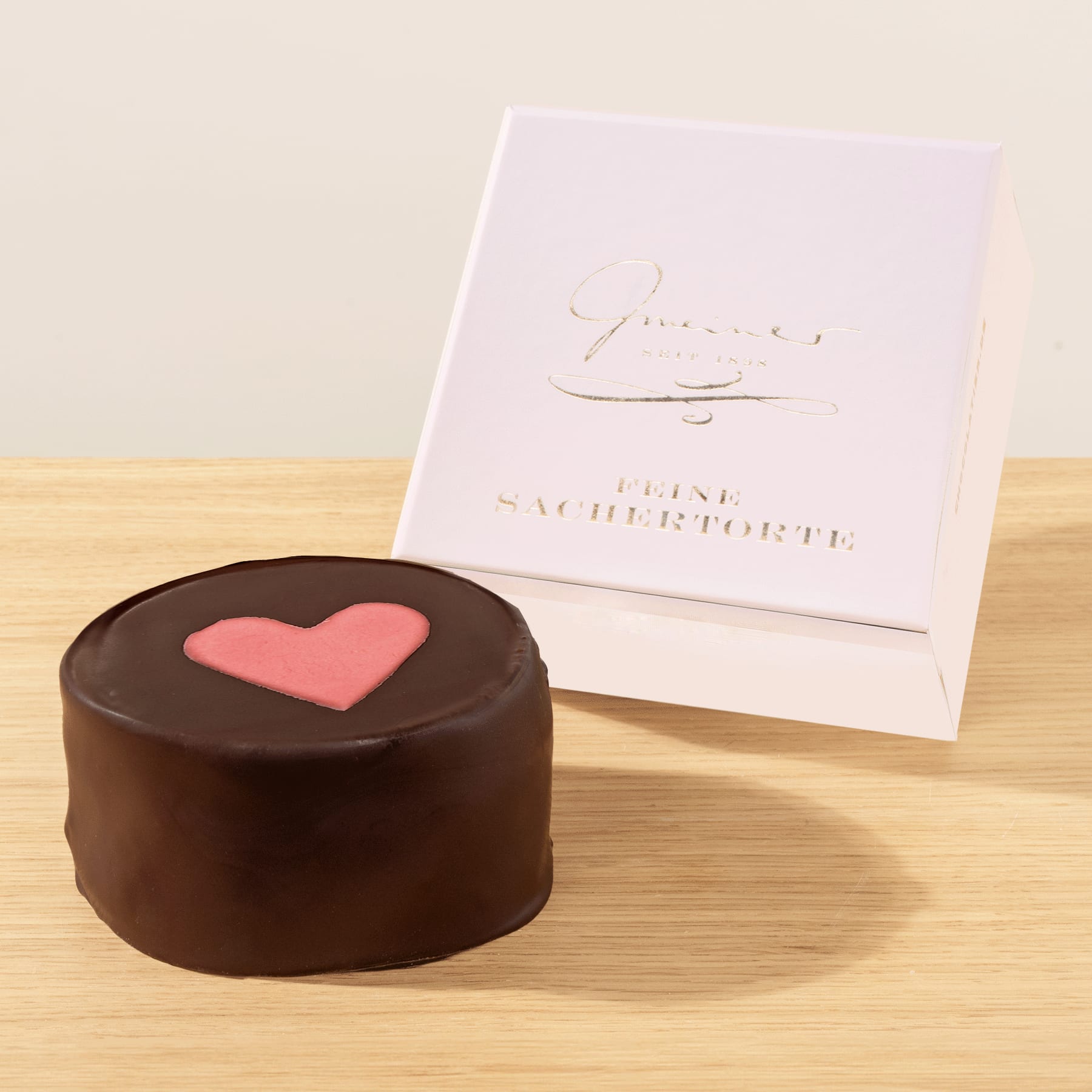 Auf einer Holzoberfläche liegt ein mit Schokolade überzogenes Dessert mit einem rosafarbenen herzförmigen Design, begleitet von einer weißen Karte mit kursivem Text.