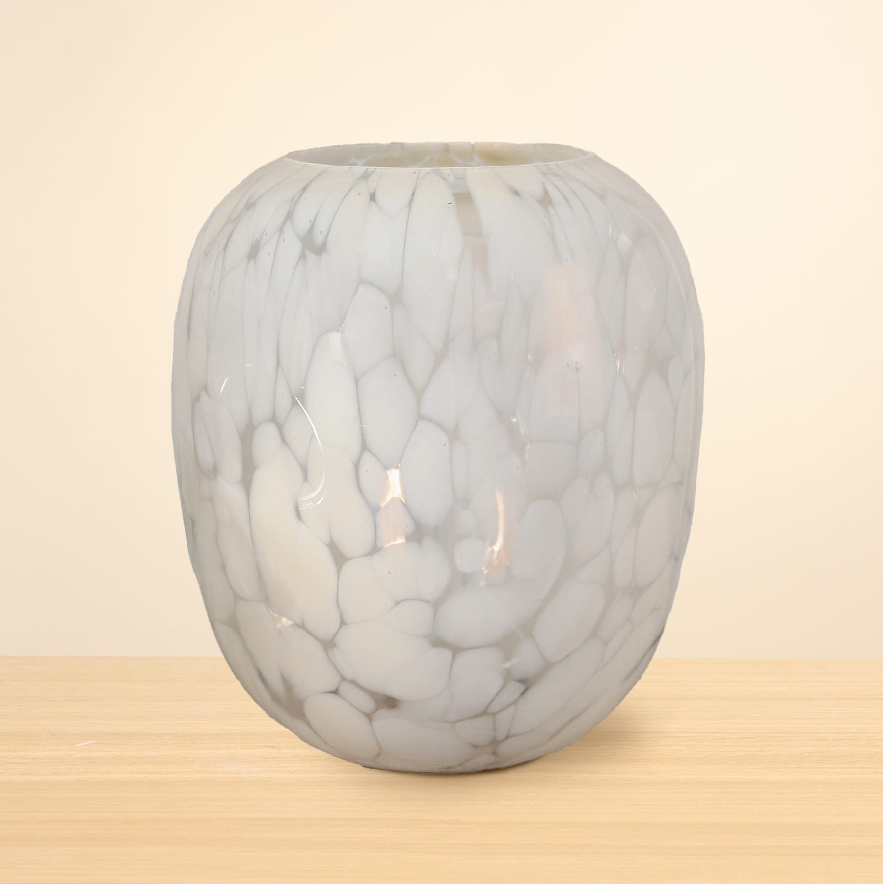 Eine runde Glasvase mit einem marmorierten, weiß-grauen Muster steht auf einer Holzoberfläche vor einem schlichten, beigen Hintergrund.