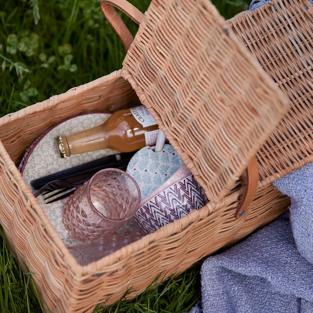 Auf einem grasbewachsenen Hintergrund steht ein geflochtener Picknickkorb, gefüllt mit verschiedenen Gegenständen, darunter eine Flasche, ein Hut und eine Decke.