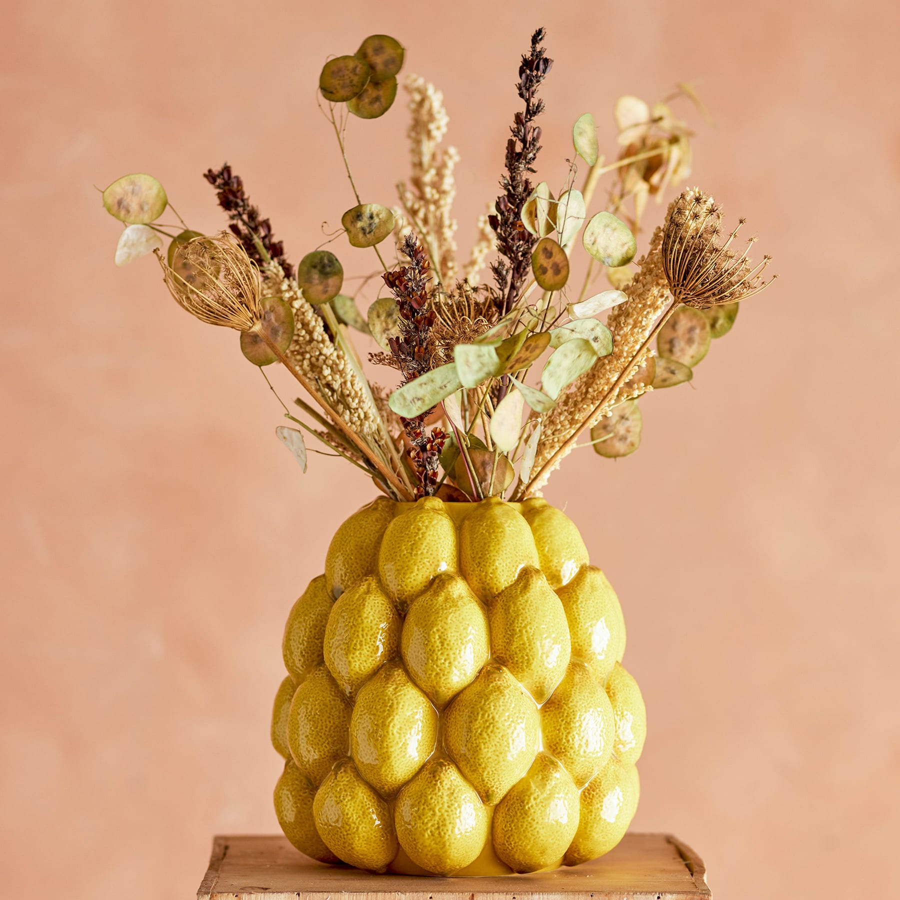 Ein farbenfroher Strauß getrockneter Blumen und Schoten in einer auffälligen gelben Vase in Ananasform steht vor einem warmen pfirsichfarbenen Hintergrund.