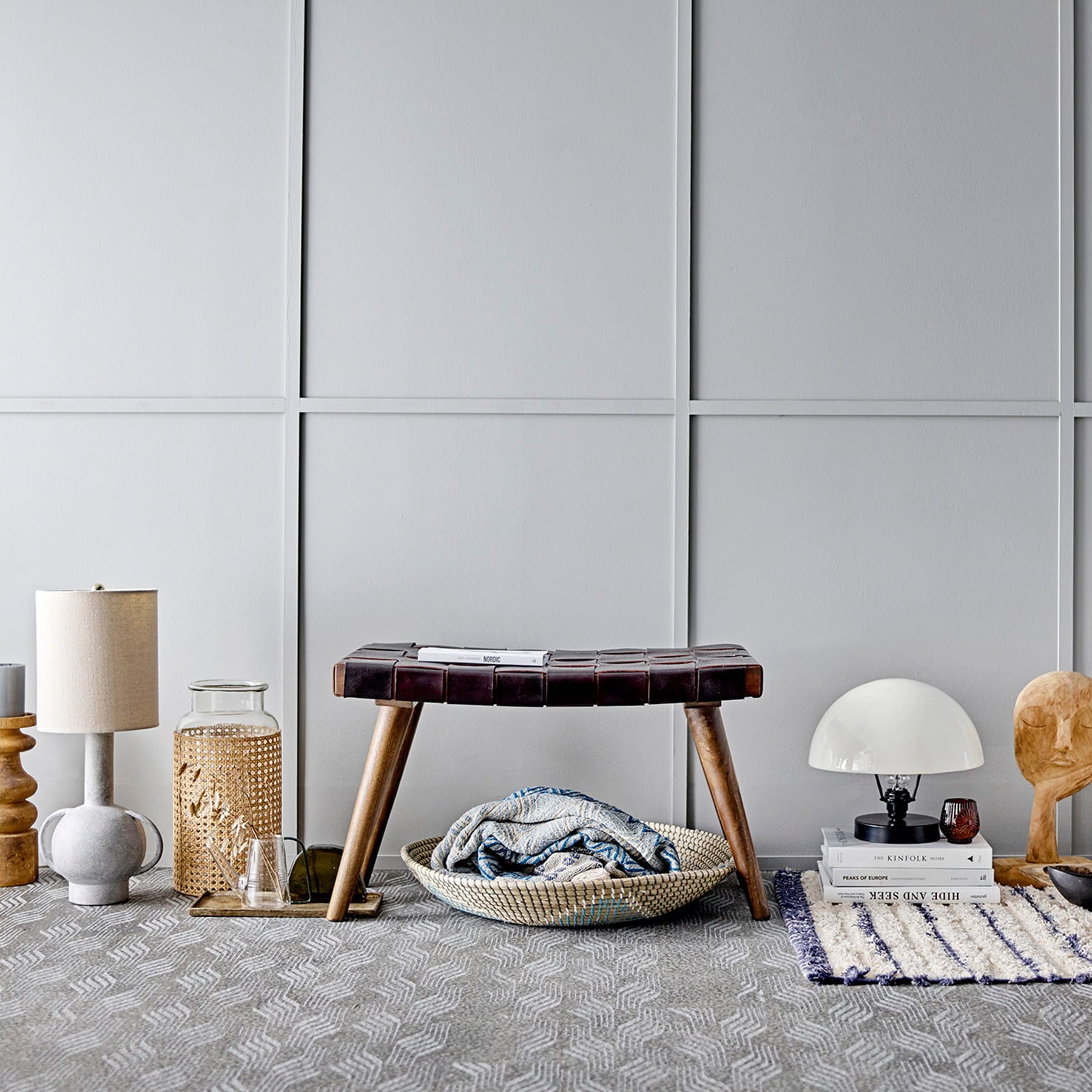 Eine gemütliche Wohnzimmereinrichtung mit einem Holzhocker, dekorativen Lampen und einem gemusterten Teppich auf dem Boden, alles vor einer minimalistischen grauen Wandkulisse.