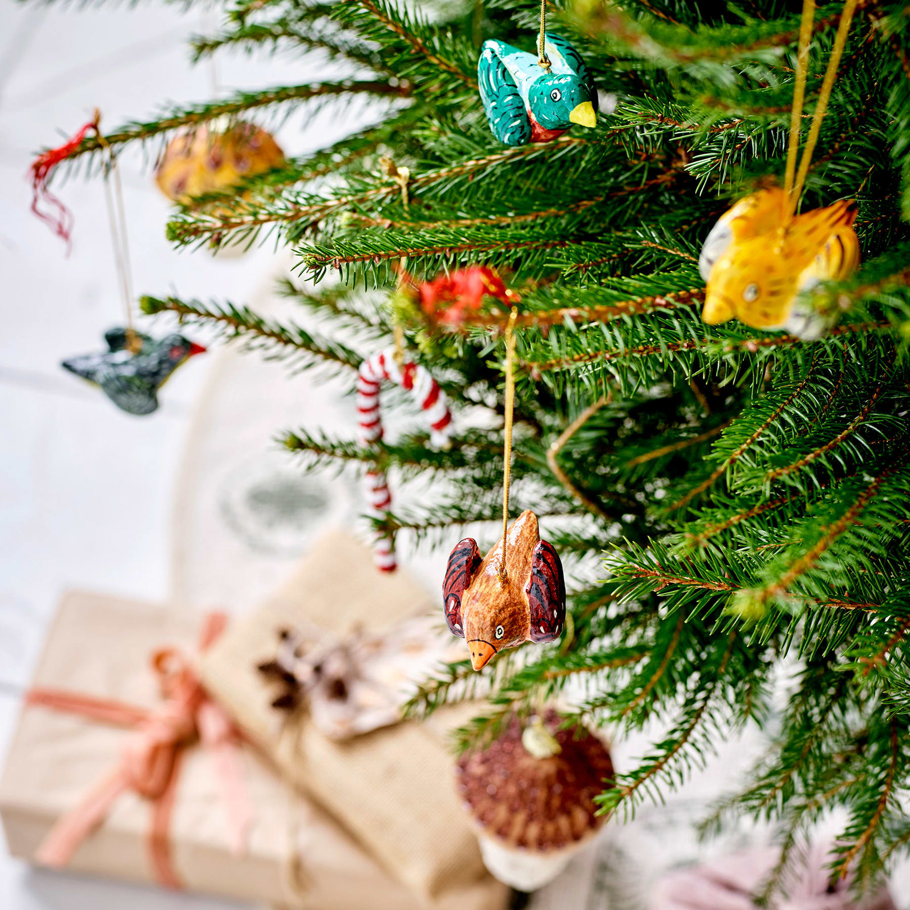 Ein festlich geschmückter Weihnachtsbaum mit verschiedenen Ornamenten, darunter ein kleiner blauer Vogel, Zuckerstangen und verpackte Geschenke im Vordergrund, vor einem unscharfen Hintergrund.