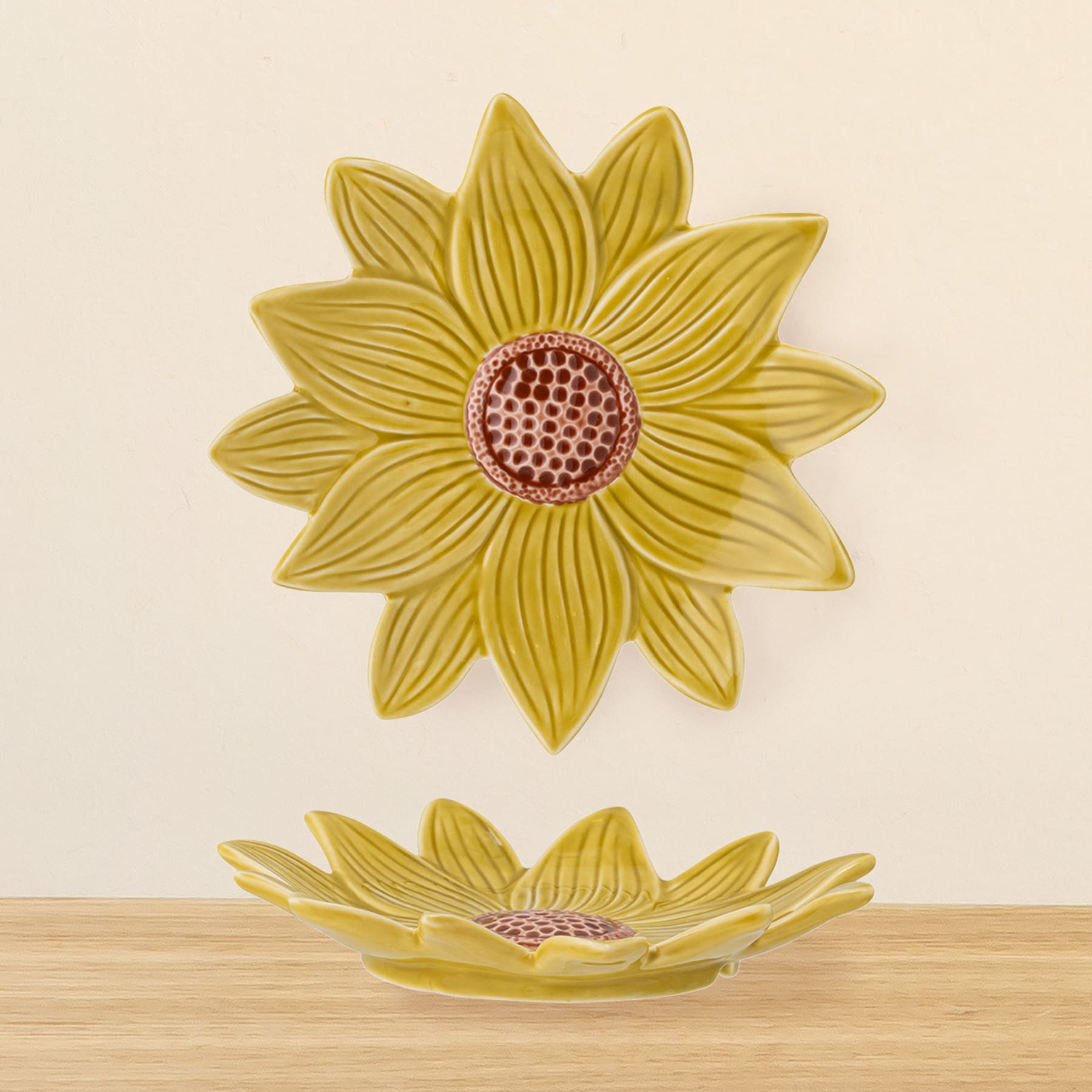 Eine große, leuchtend gelbe Keramikschale in Form einer Sonnenblume steht auf einer Holzoberfläche. Ihre kunstvollen Blütenblätter und der zentrale, rot gepunktete Kern bilden einen auffälligen optischen Kontrast zum schlichten Hintergrund.