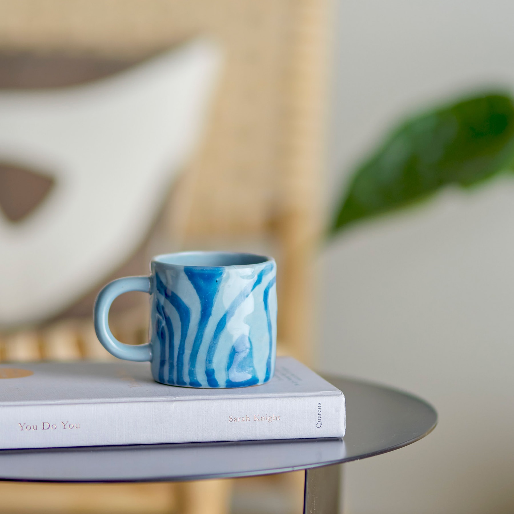 Auf einem Stapel Bücher steht eine blau-weiß marmorierte Keramiktasse, im Hintergrund ist ein grünes Pflanzenblatt zu sehen.