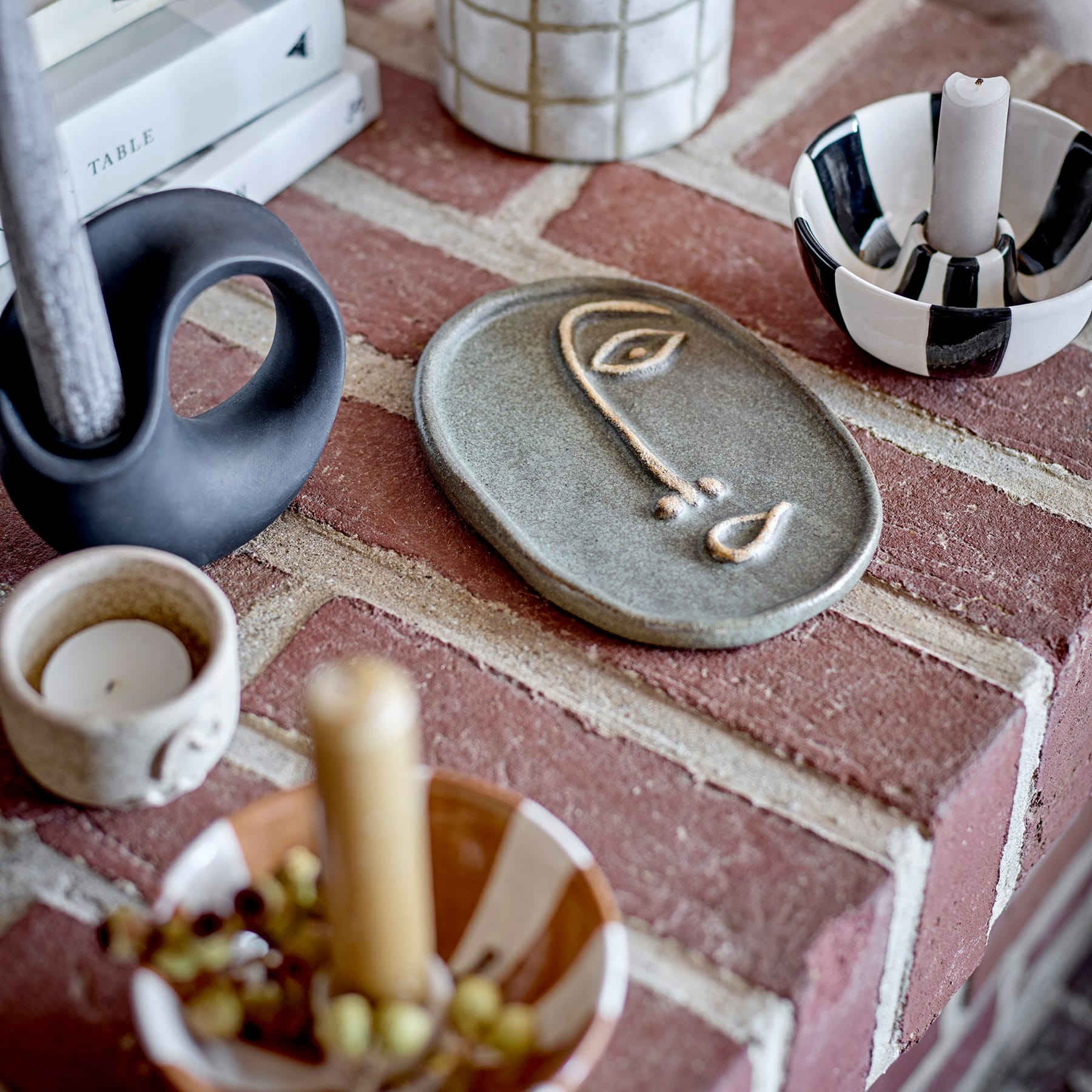 Auf einer Ziegeloberfläche steht ein keramisches Objekt in Form eines Gesichts, umgeben von verschiedenen Kunstgegenständen und Materialien, darunter eine Kerze, eine Tasse und ein Stifthalter.