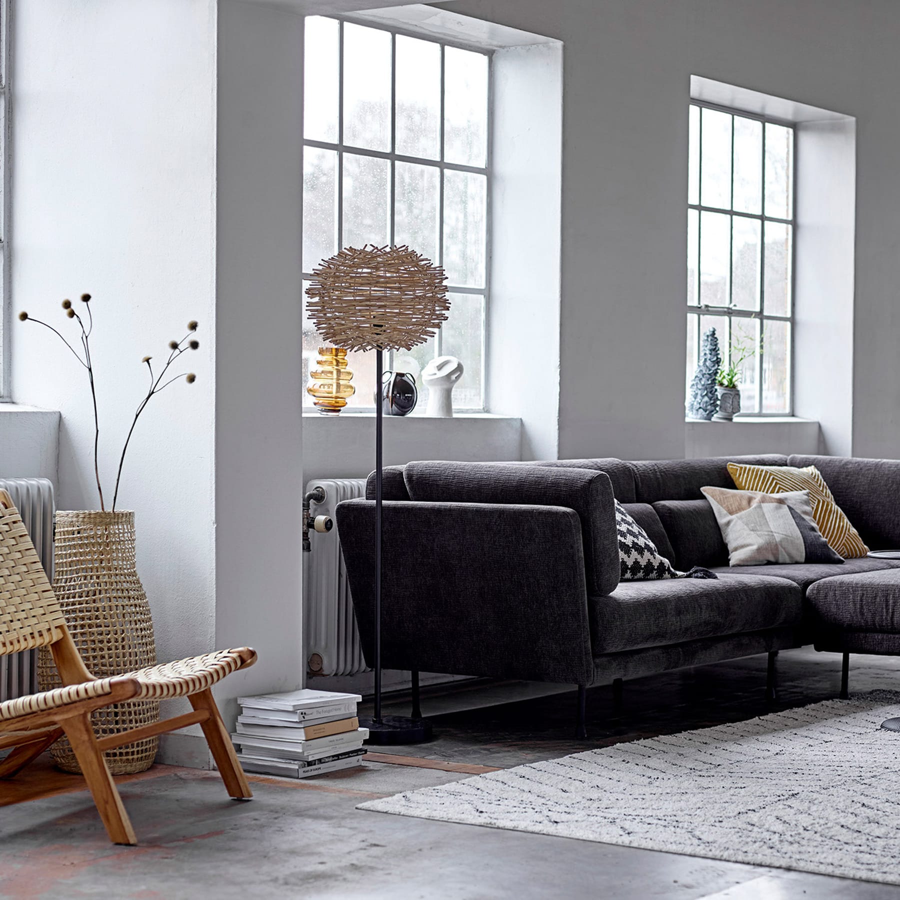 Ein gemütliches und modernes Wohnzimmer mit einem großen schwarzen Sofa, einem geflochtenen Sessel und einer einzigartigen Leuchte aus geflochtenen Zweigen vor dem Hintergrund großer Fenster mit Blick auf eine ruhige Außenszene.