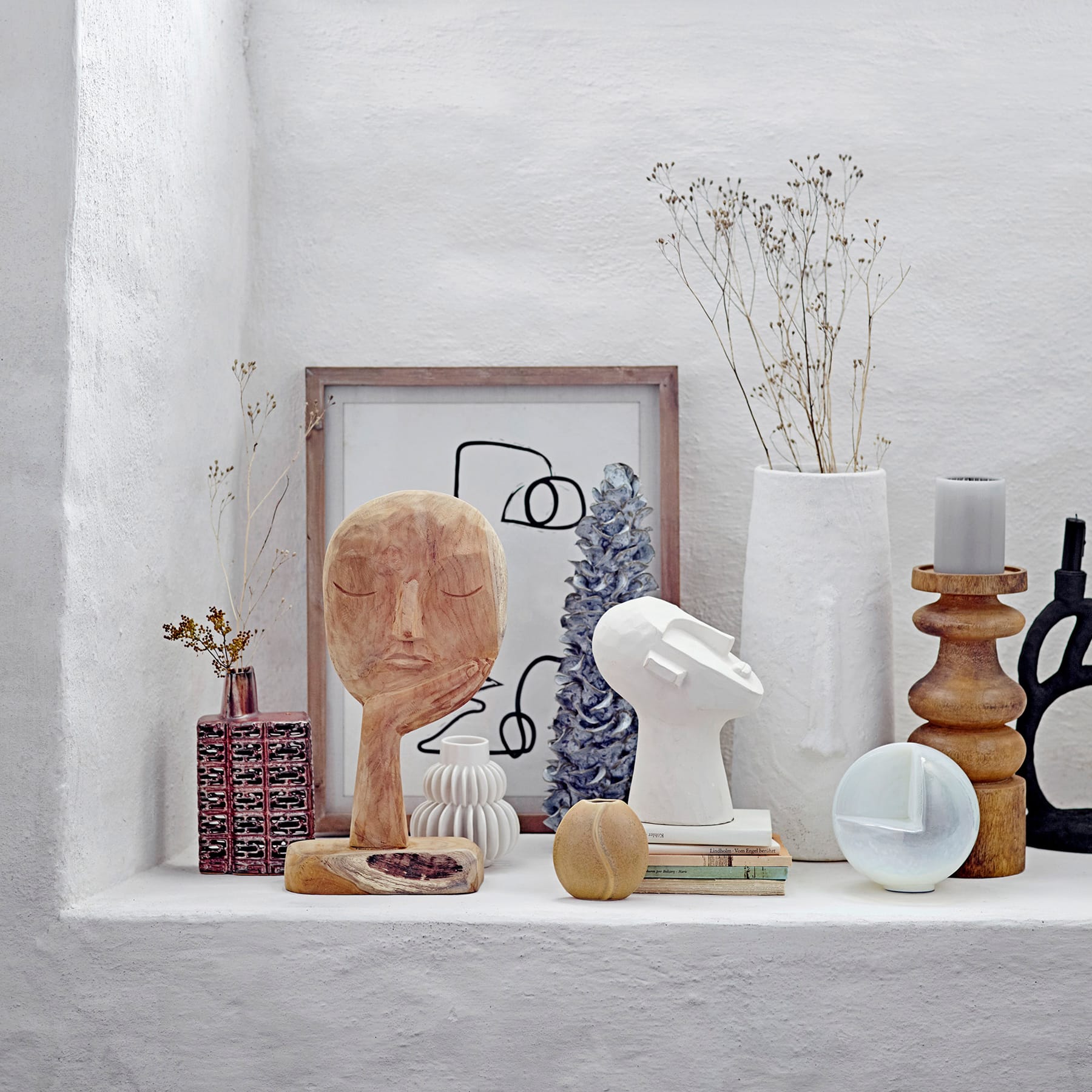 Ein minimalistisches Stillleben-Arrangement mit verschiedenen abstrakten Skulpturen, gerahmten Kunstwerken und Dekorationsobjekten auf einer weißen Oberfläche vor einem schlichten Hintergrund.