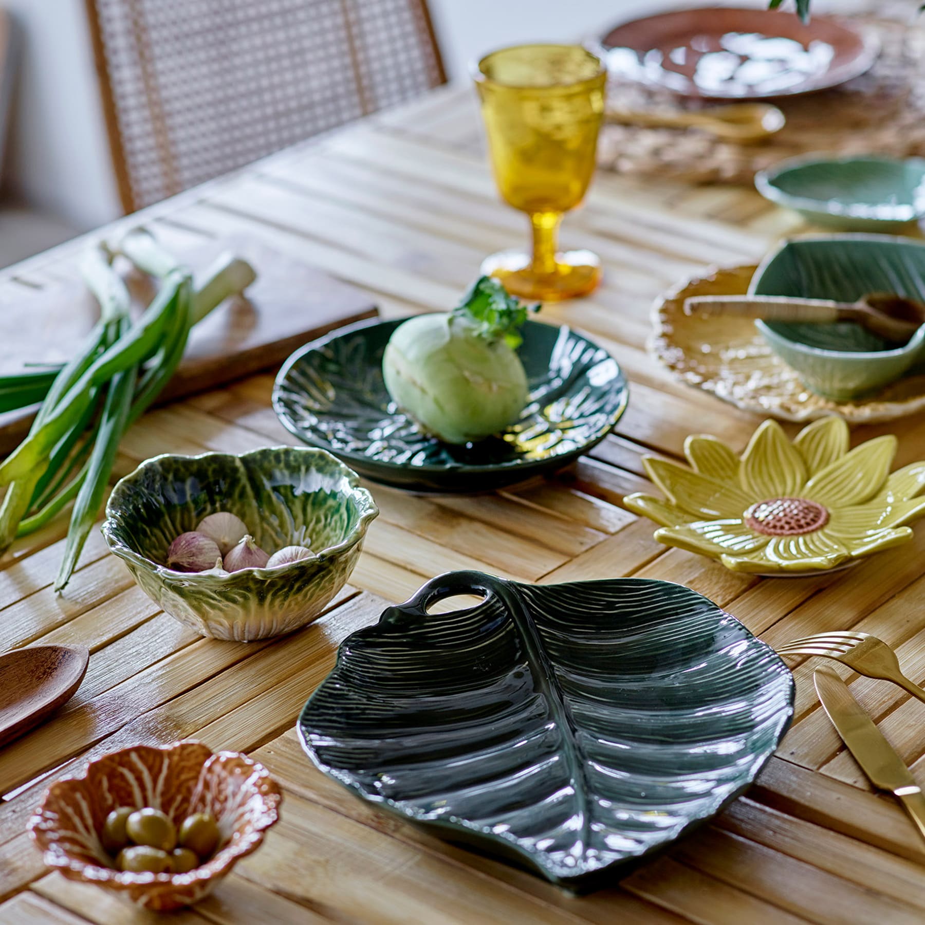 Ein Holztischset mit verschiedenen Keramikgeschirren, darunter ein blattförmiger Teller, ein grüner Apfel und ein gelbes Glas, umgeben von dekorativen Blumenelementen.