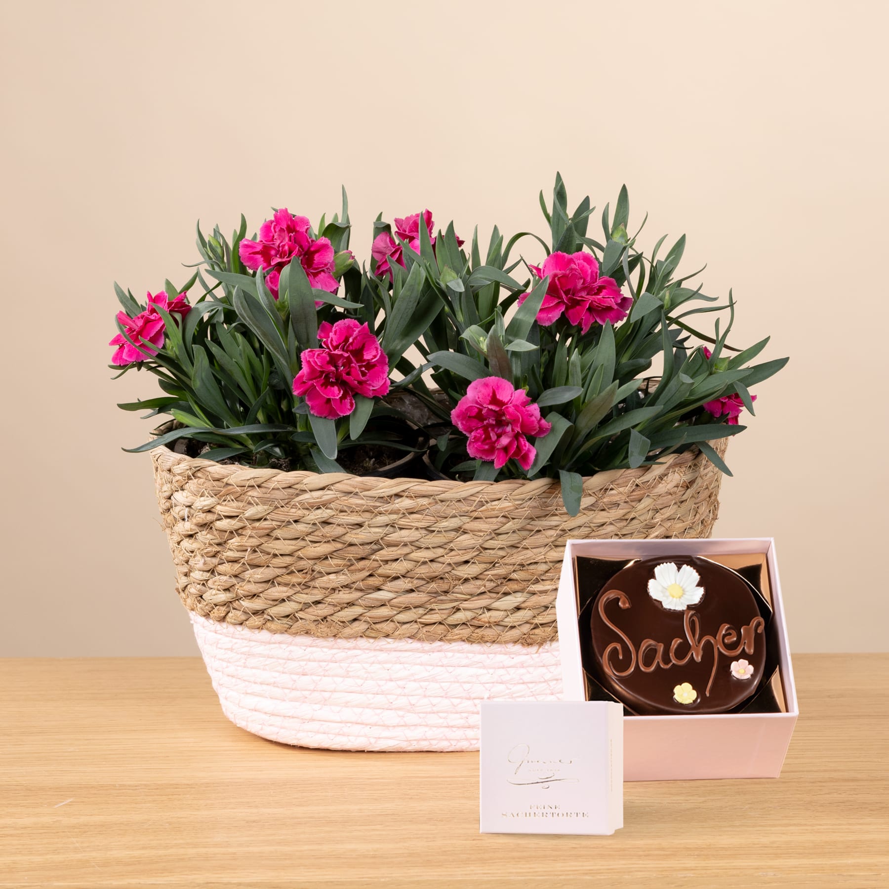 Auf einem Holztisch steht ein geflochtener Korb voller leuchtend rosa Nelkenblüten, daneben eine kleine Pralinenschachtel mit der Aufschrift „Sachet“.