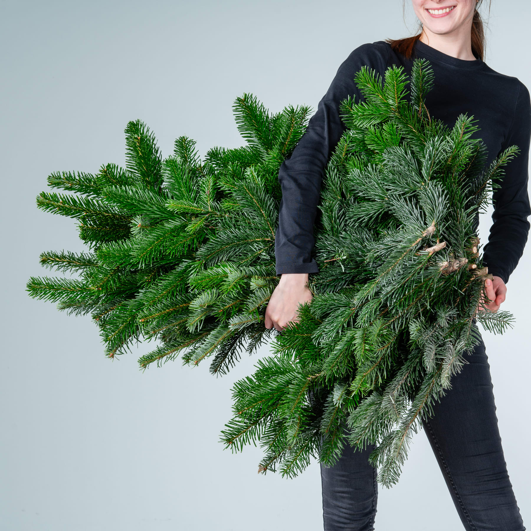 Eine Person in einem schwarzen Pullover hält einen großen, üppigen und leuchtenden immergrünen Kranz oder ein Bündel Tannenzweige vor einem schlichten, hellen Hintergrund.